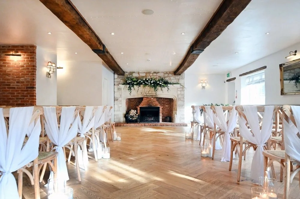Elegant wedding or event venue with rows of wooden chairs draped with white fabric, a brick and stone fireplace decorated with greenery, and candles on the floor, inside a bright room with wooden beams and large windows.