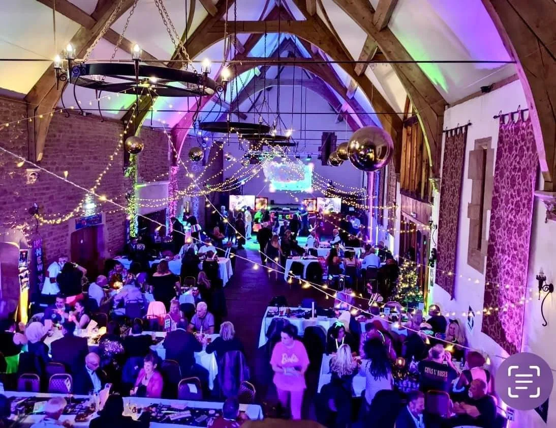 Indoor event in a large hall with wooden beams, brick walls, and colorful lighting. Guests are gathered around tables, dancing, and socializing. String lights, disco balls, and a stage with a large screen create a festive atmosphere.