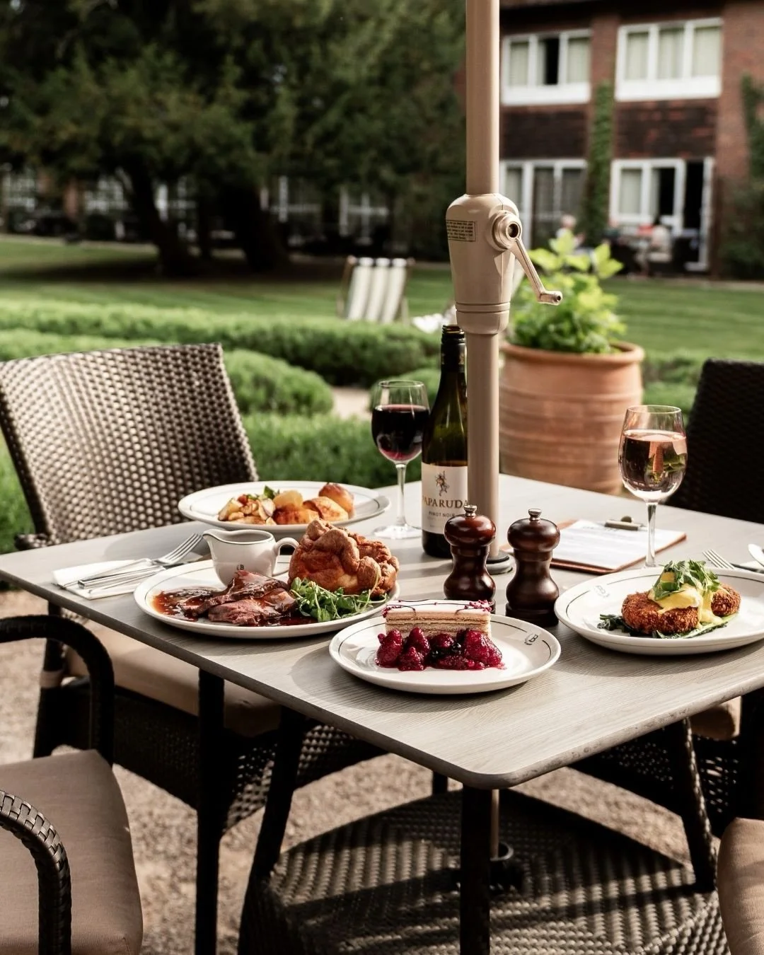 Outdoor dining table set with plates of meat, fried chicken, cheesecake, and berry compote, along with glasses of red wine, salt and pepper shakers, a bottle of wine, and surrounded by wicker chairs, in a garden setting.