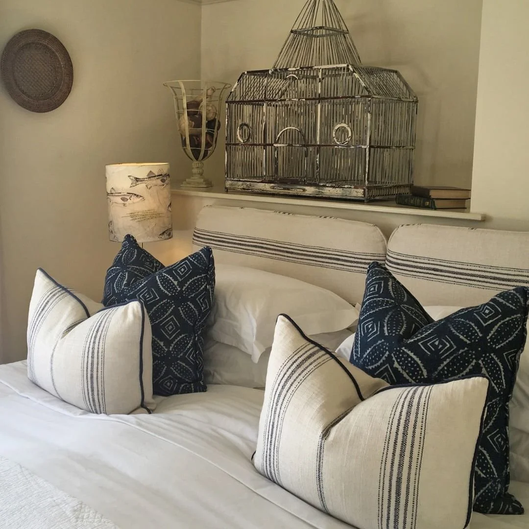 A cozy bedroom with a neatly made bed, multiple decorative pillows in navy and white patterns, a fabric lamp with a fish design, and a vintage birdcage decor piece on the shelf above the bed. Additional books and decorative items are also on the shel