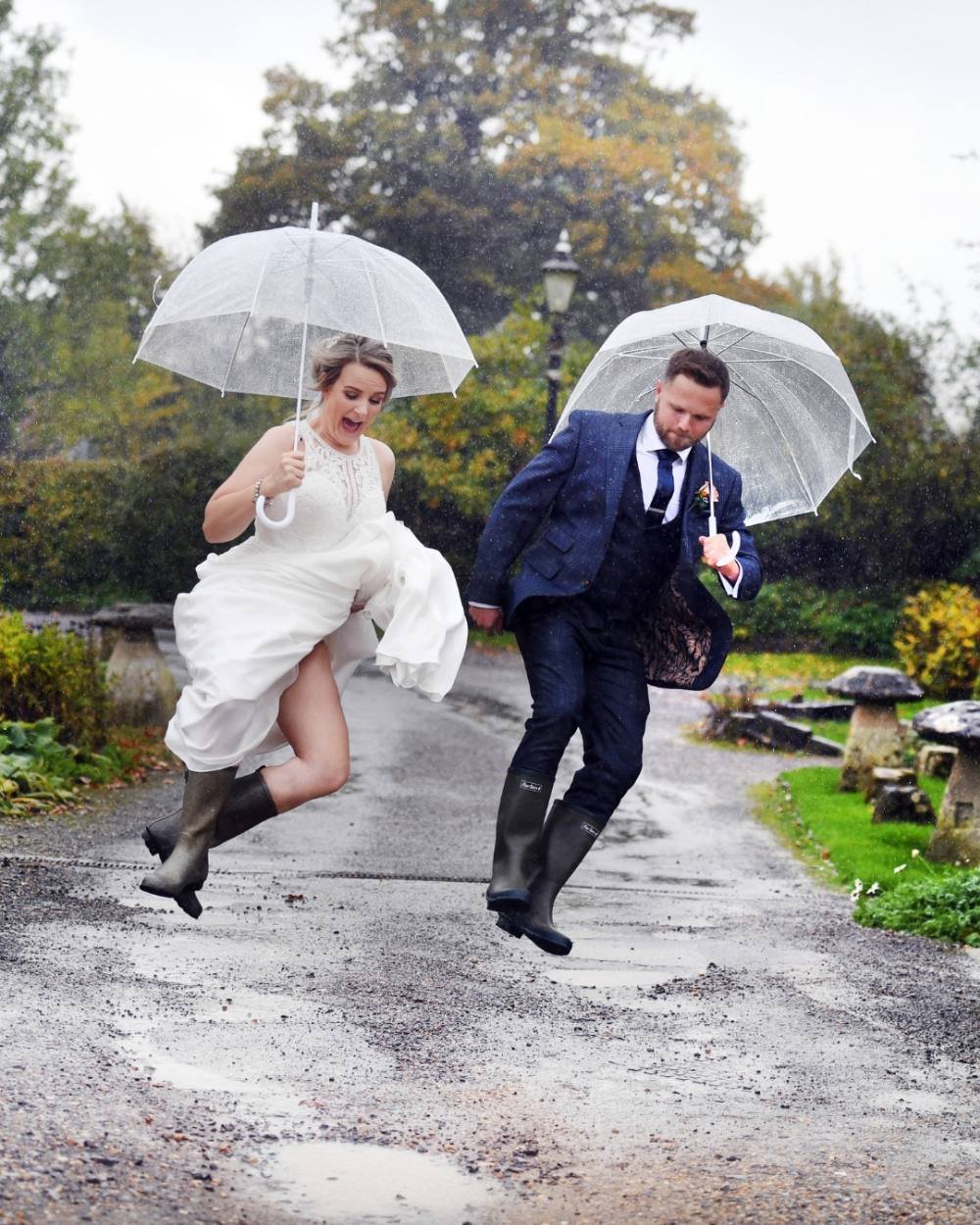 A couple in wedding attire jumping over puddles in the rain, holding clear umbrellas, on a garden pathway.