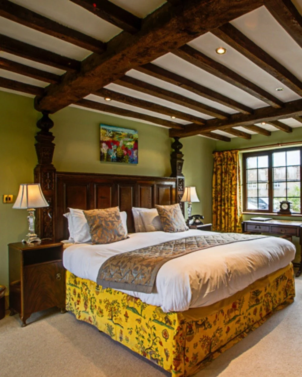 A bedroom with a large bed, decorative pillows, wooden headboard, and matching side tables with lamps, featuring a window with floral curtains and exposed wooden beam ceiling, in a cozy, traditional style.