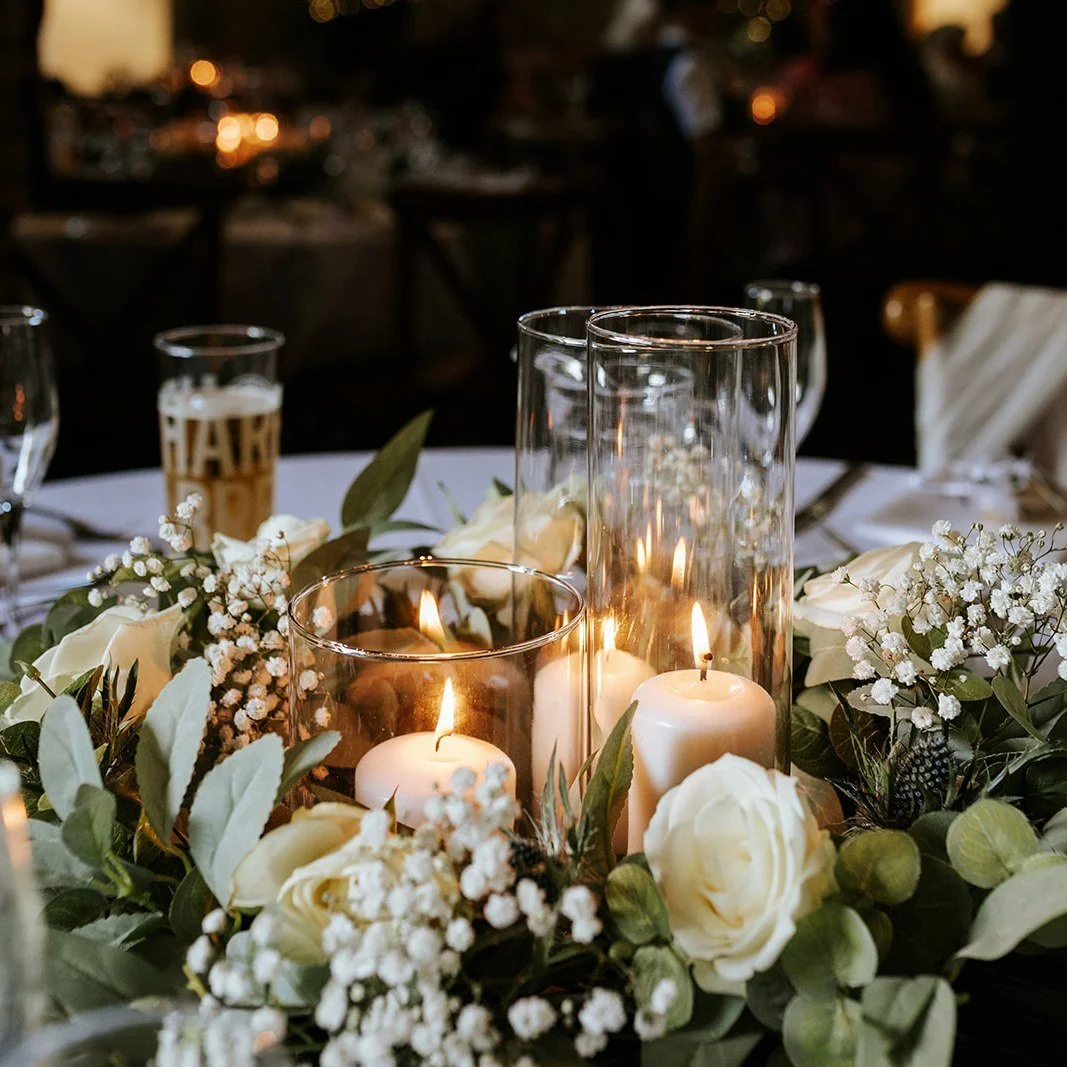 A table centerpiece at a formal event with white roses, baby's breath, green leaves, candles in glass holders, and a drink with a golden colored straw in the background.