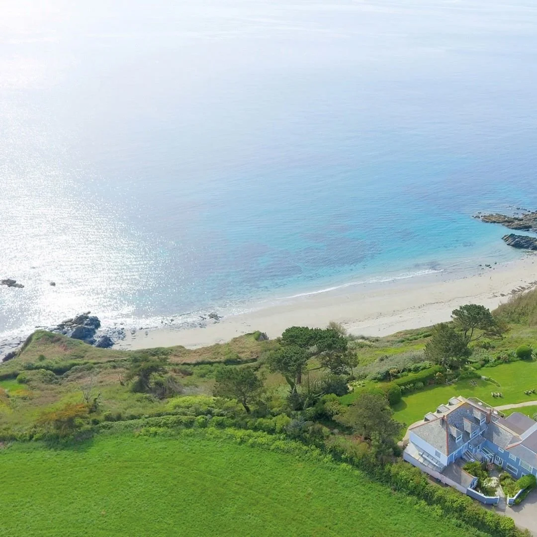 Aerial view of a coastal landscape with a sandy beach, green grassy areas, trees, and a house near the shoreline with a driveway.