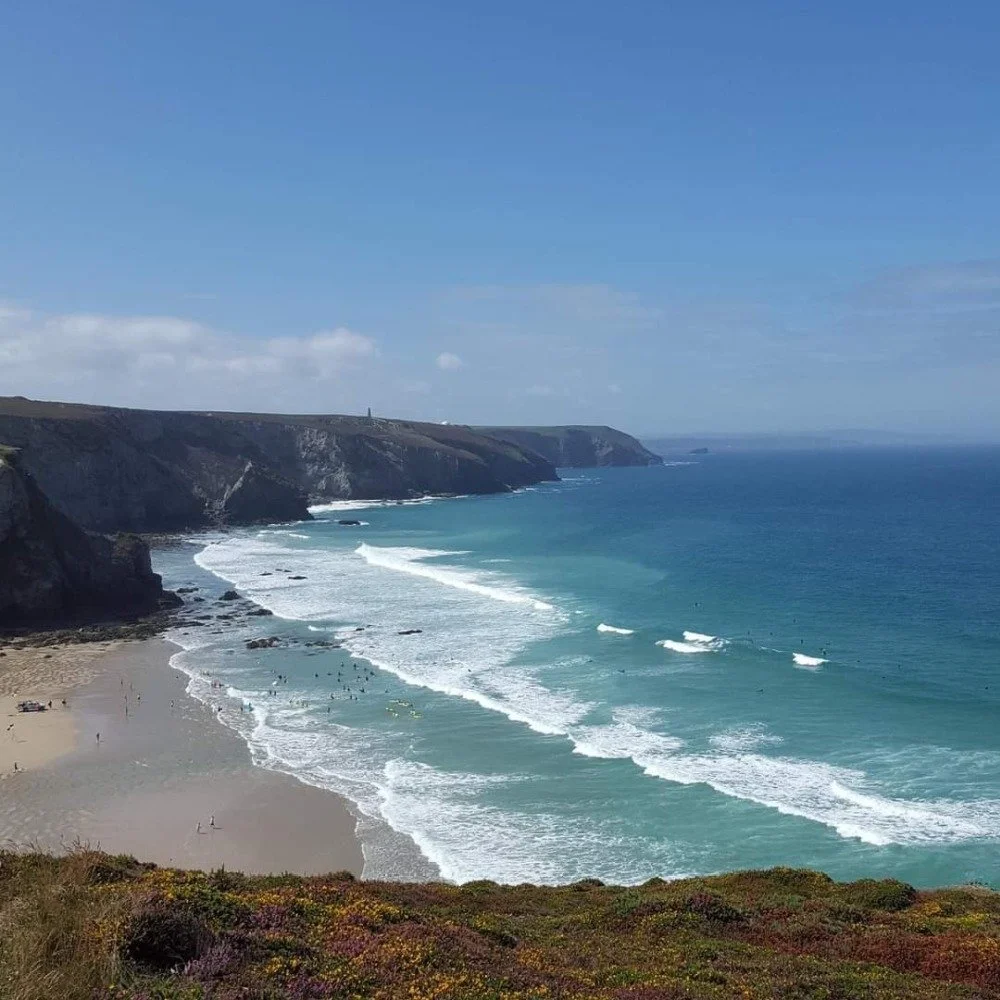 Scenic view of a coastline with cliffs on the left, sandy beach below, and ocean waves crashing onto the shore. Blue sky with some clouds.