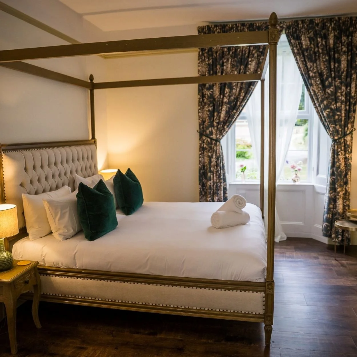 A cozy bedroom with a large four-poster bed, white bedding, and dark green pillows. Two rolled towels are placed on the bed. There is a wooden nightstand with a lamp on each side of the bed. The room has dark wood flooring and a window with floral cu