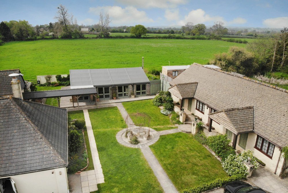 Aerial view of a houses and garden with green lawn, trees, and a field in the background.