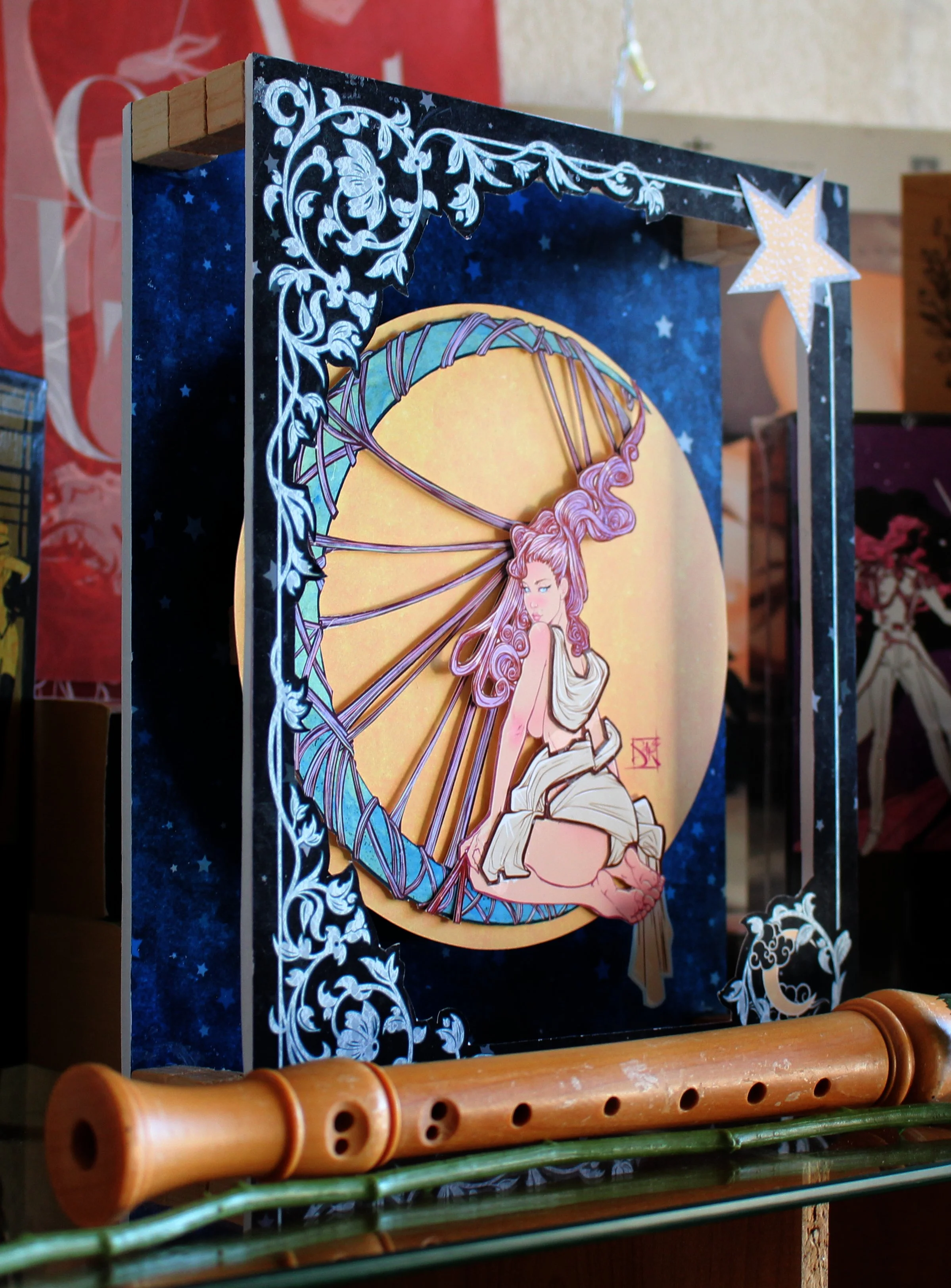 A decorative display featuring a framed artwork of a woman with long, flowing hair, sitting with her knees drawn up, surrounded by a starry night sky, with a star-shaped ornament hanging from the top right corner of the frame.