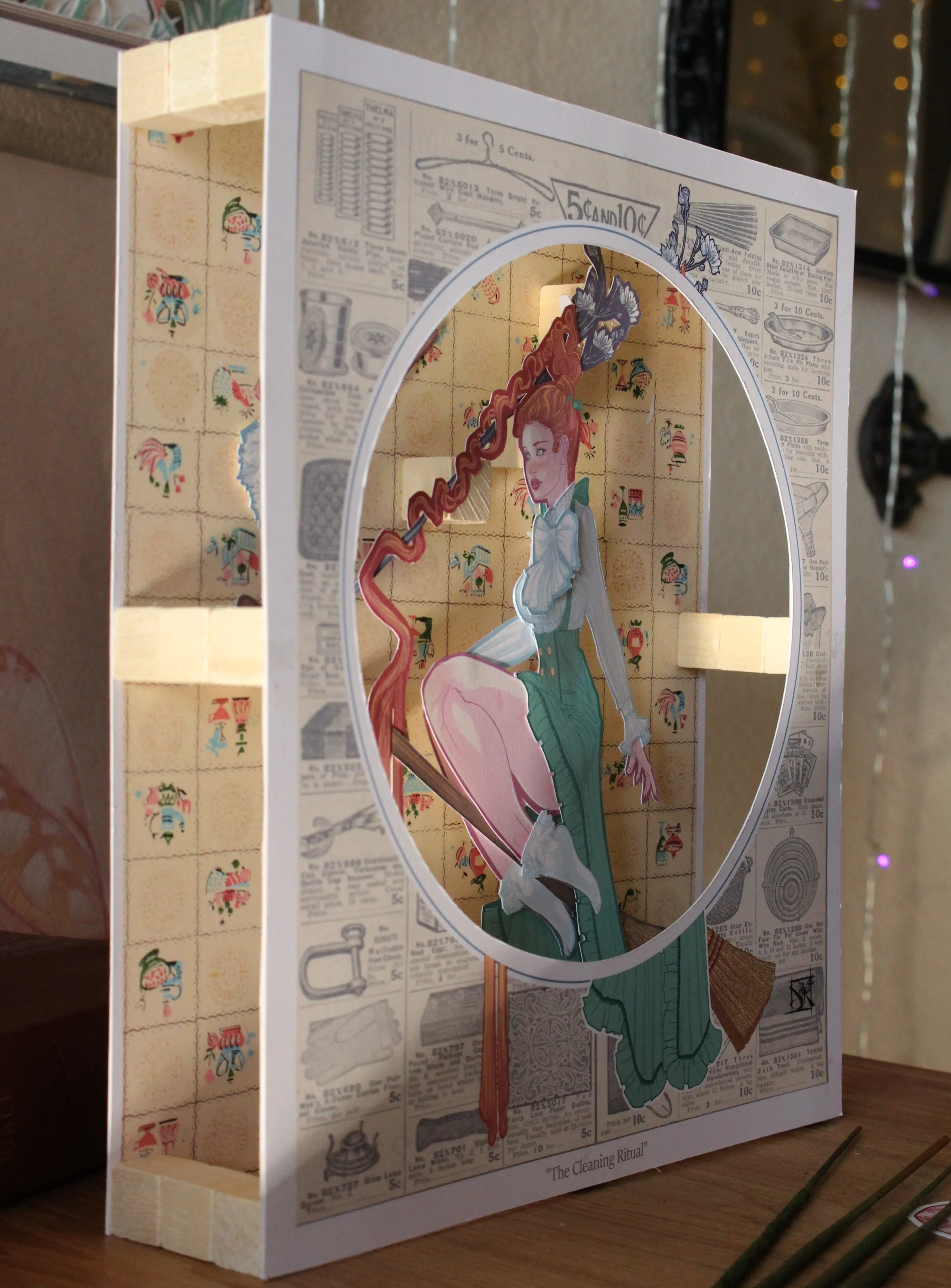 A shadow box with vintage newspaper clippings on the sides and a scene with a woman with red hair sitting on a chair, wearing a green dress, in the center. The shadow box is titled 'The Cleaning Ritual' at the bottom.