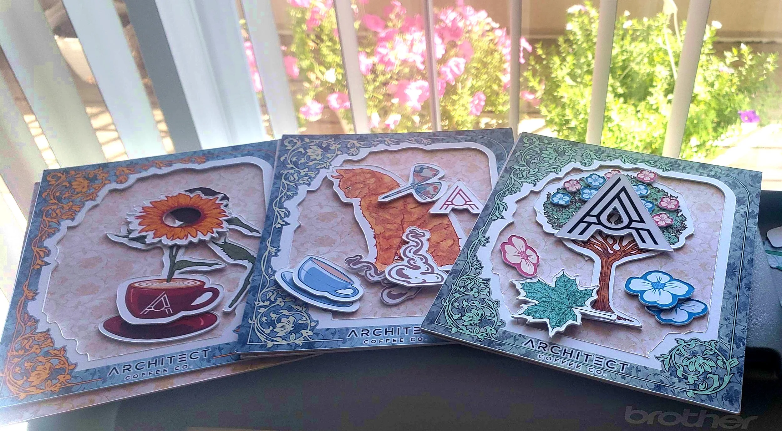 Set of colorful 3D mini-boxes prior to assembly from artist Anthony Christopher Robinson, depicting a sunflower in a cup, a curious kitty, and a tree with flowers, laid out on the printer with a window and flowers in the background.