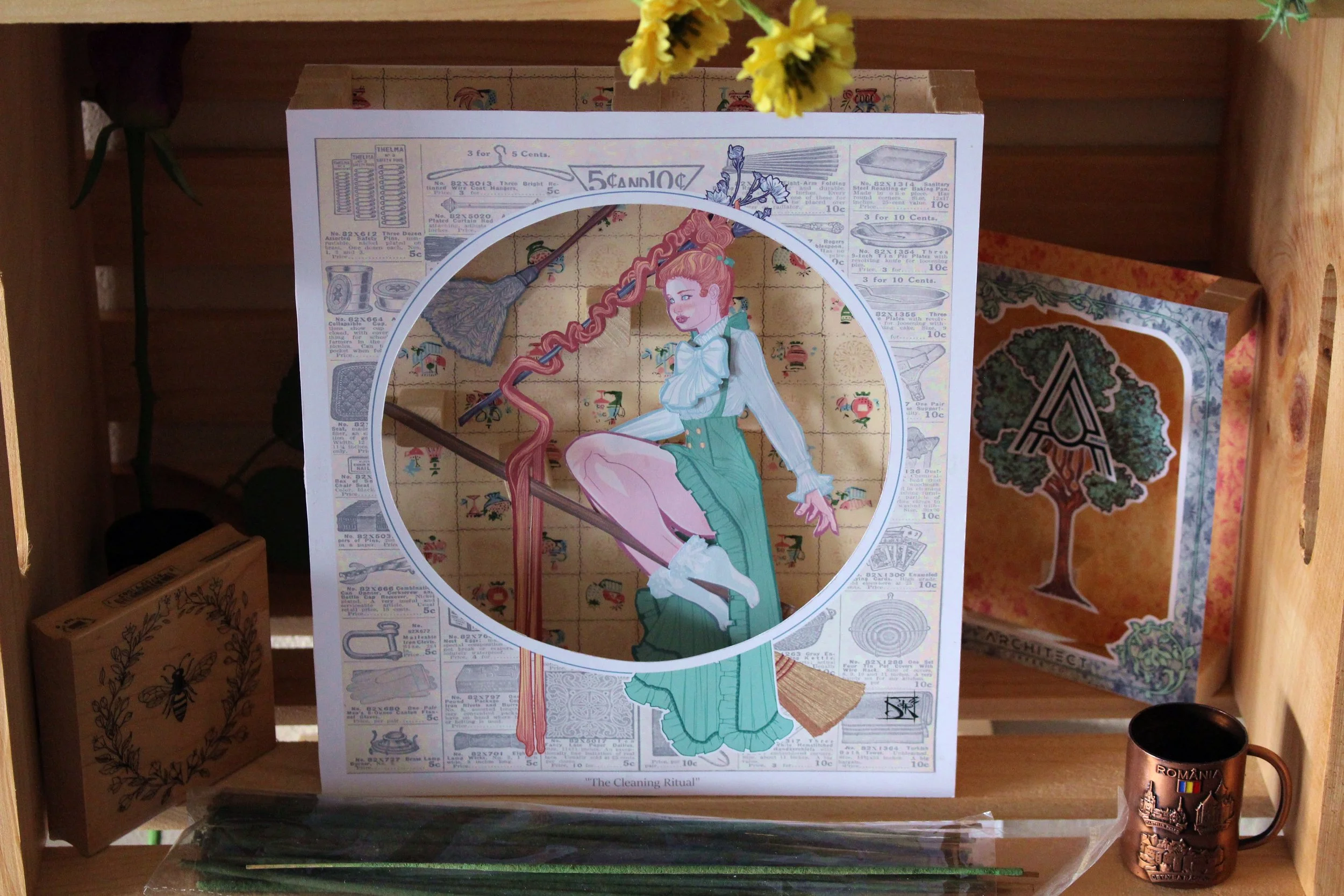 A shelf displaying a vintage-style illustrated card titled 'The Cleaning Ritual' with a woman in a green skirt and white blouse holding a mop. Surrounding the card are boxes and other decorative items, including a mug and a plant.