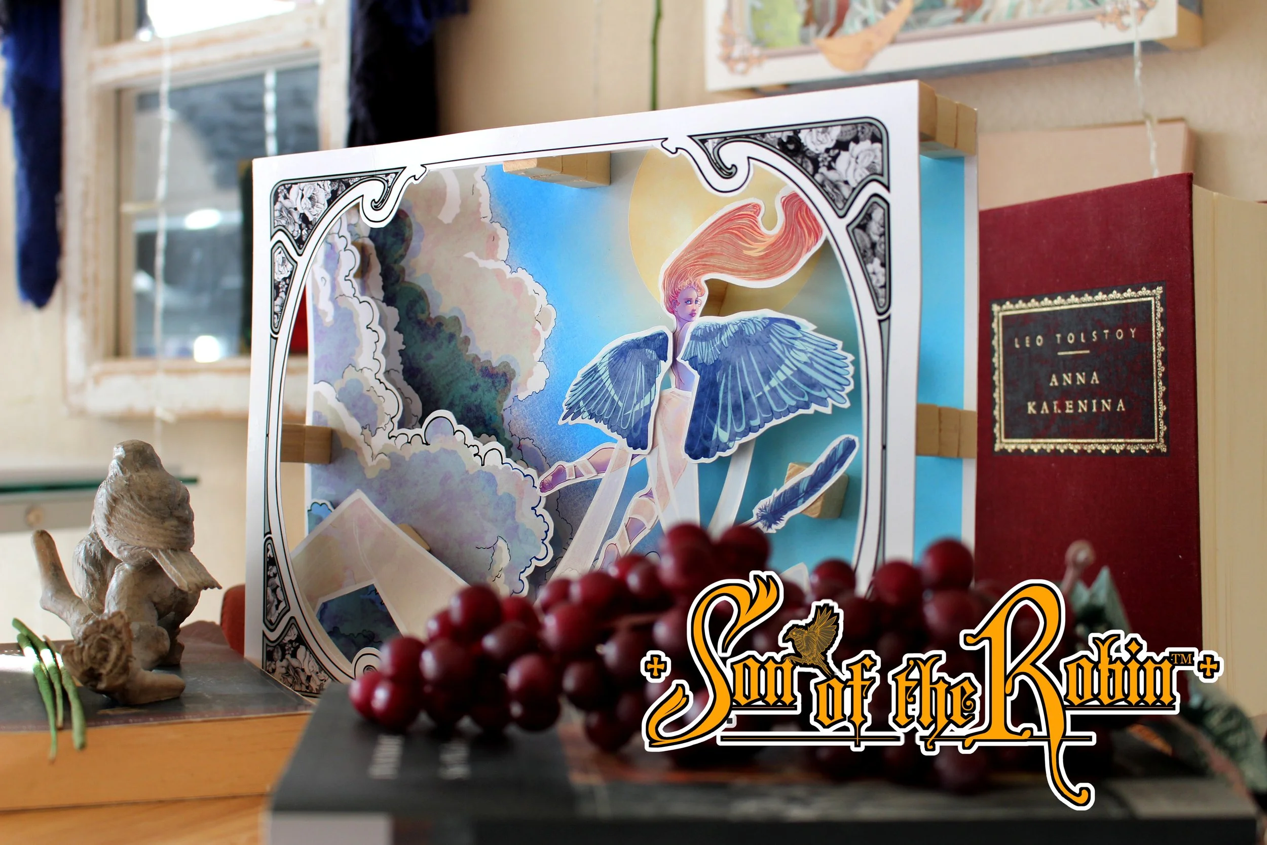 "Chasing Icarus", a colorful and whimsical 3D storybox from artist Anthony Christopher Robinson depicting a beautiful woman sailing upwards towards the sun on feathered wings, is displayed on a table with various decorative items.