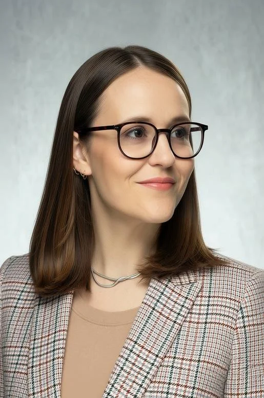 A woman with straight brown hair, wearing black-framed glasses, a beige top, plaid blazer, and a silver necklace, looking to her right with a slight smile.