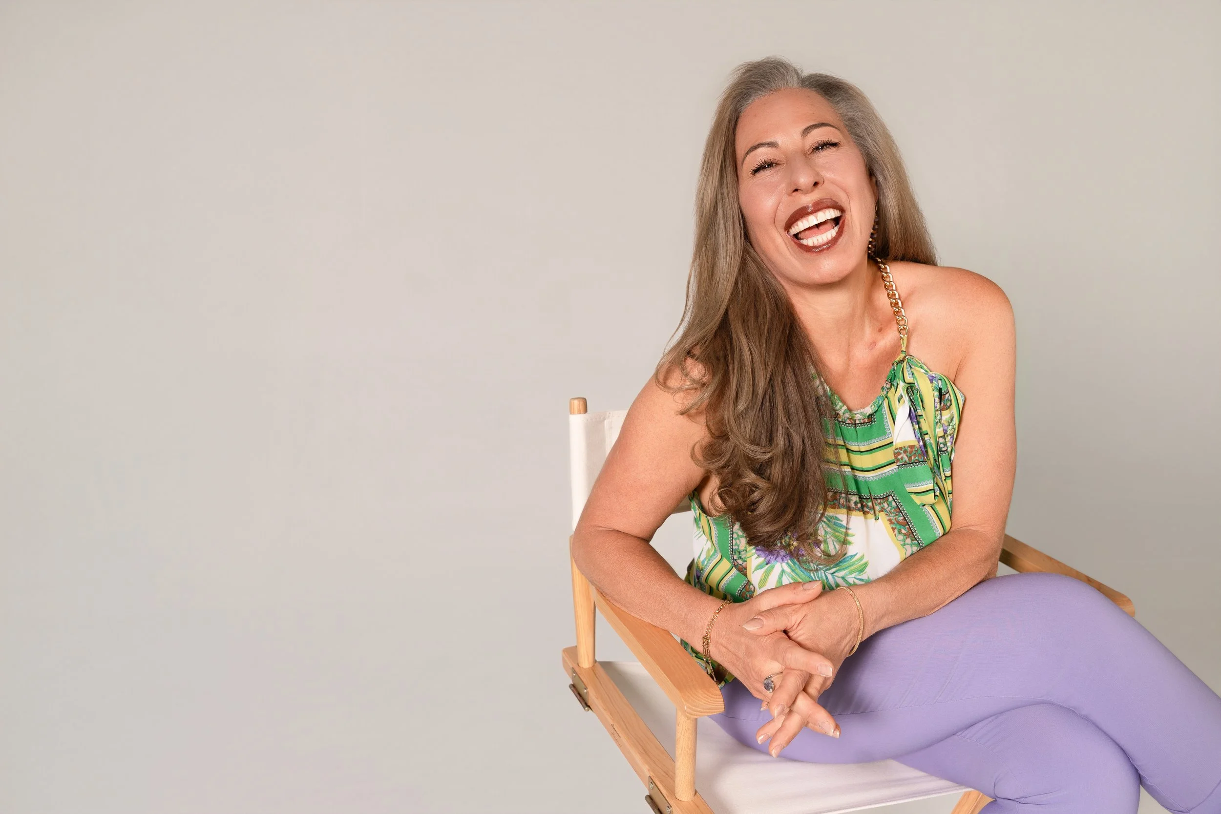 A woman with long gray hair smiling and laughing sitting on a director's chair, wearing a green patterned tank top and lavender pants, with a plain light-colored background.