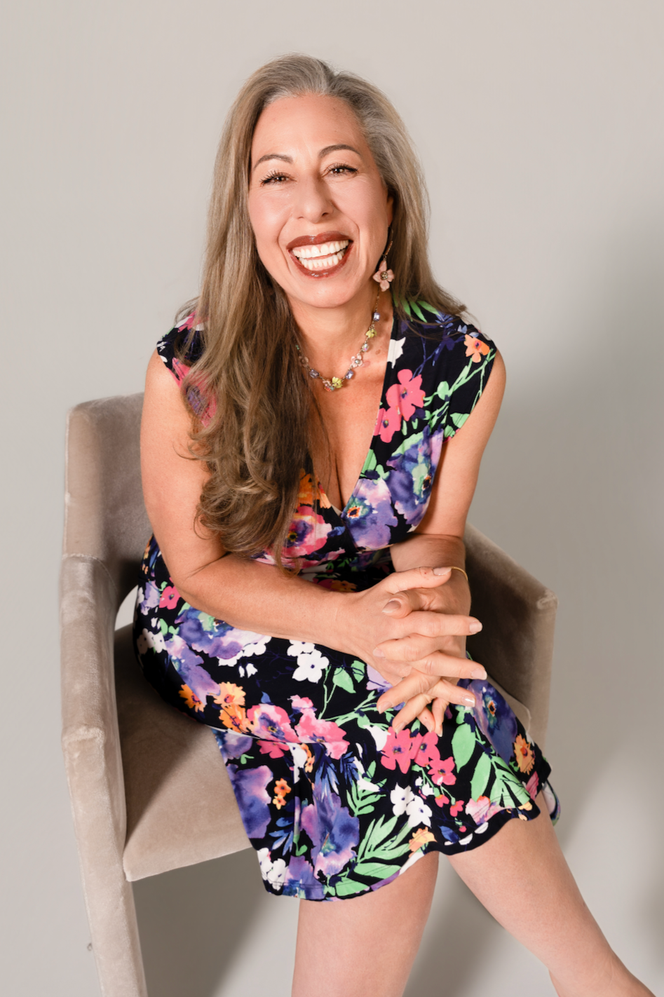 A woman with long gray hair sitting on a beige chair, smiling and wearing a colorful floral dress and jewelry.