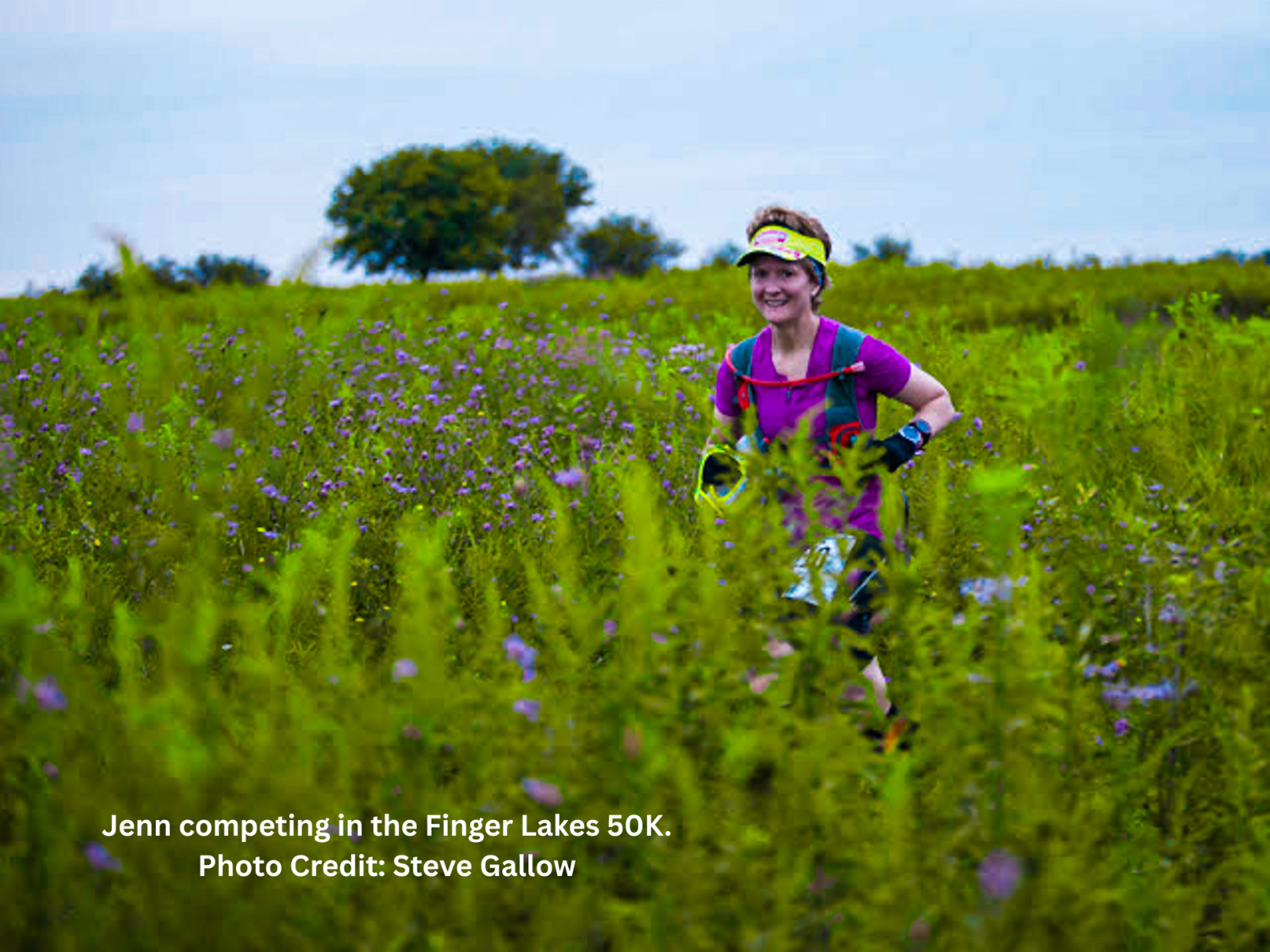 Jenn Woltjen in athletic gear, running through a field of purple flowers, during a race called the Finger Lakes 50K, with trees and a blue sky in the background.