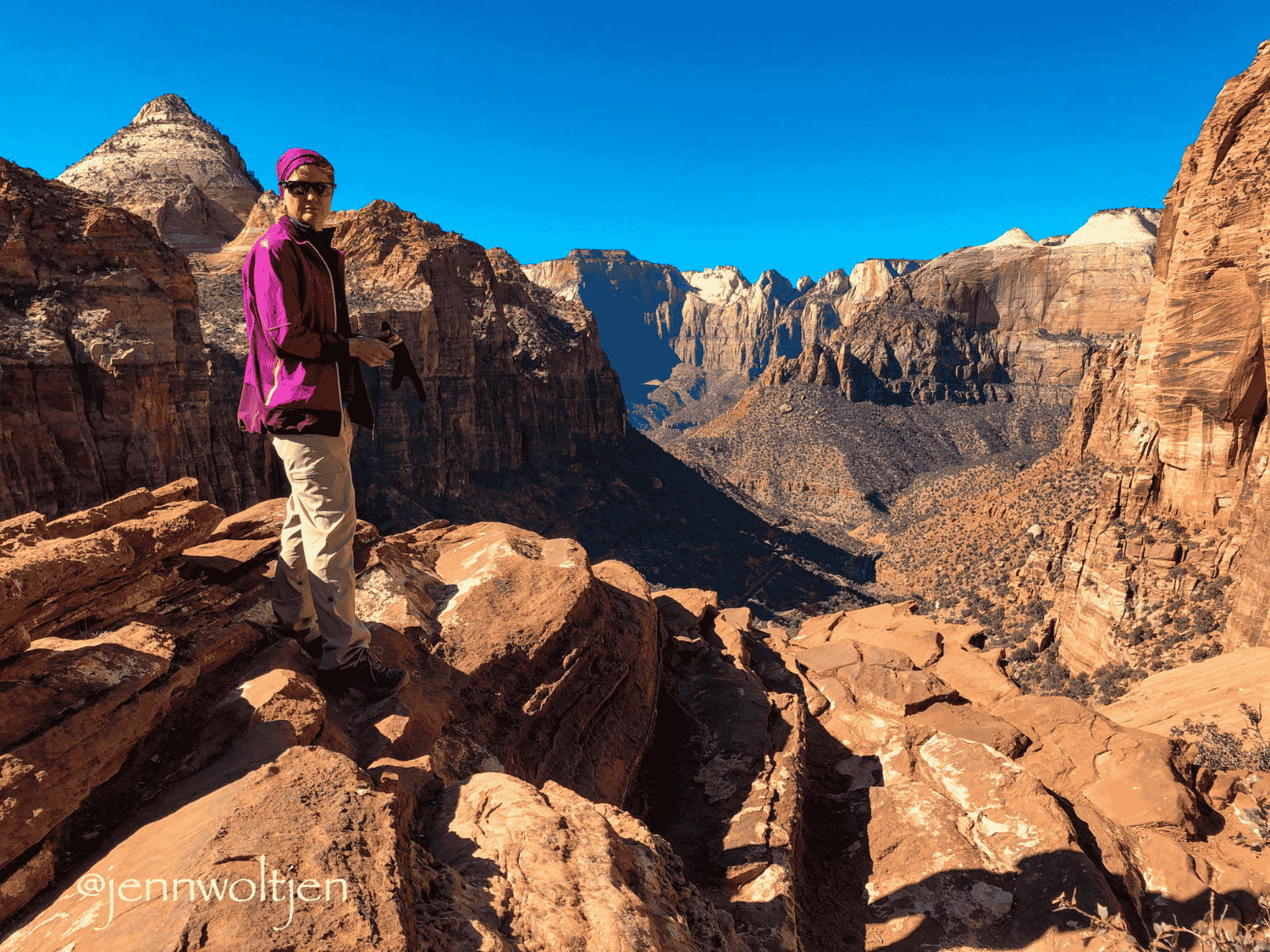 Jenn Woltjen standing on rocky terrain overlooking a deep canyon with towering cliffs and a clear blue sky.