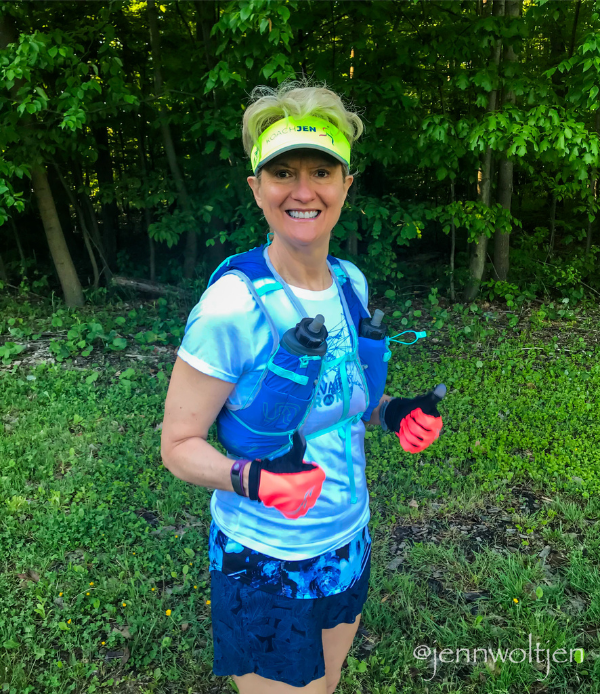 Jenn Woltjen in athletic gear standing outdoors on a grassy area with trees in the background. She is wearing a white and blue running shirt, blue shorts, a yellow headband, and a blue vest with water bottles.