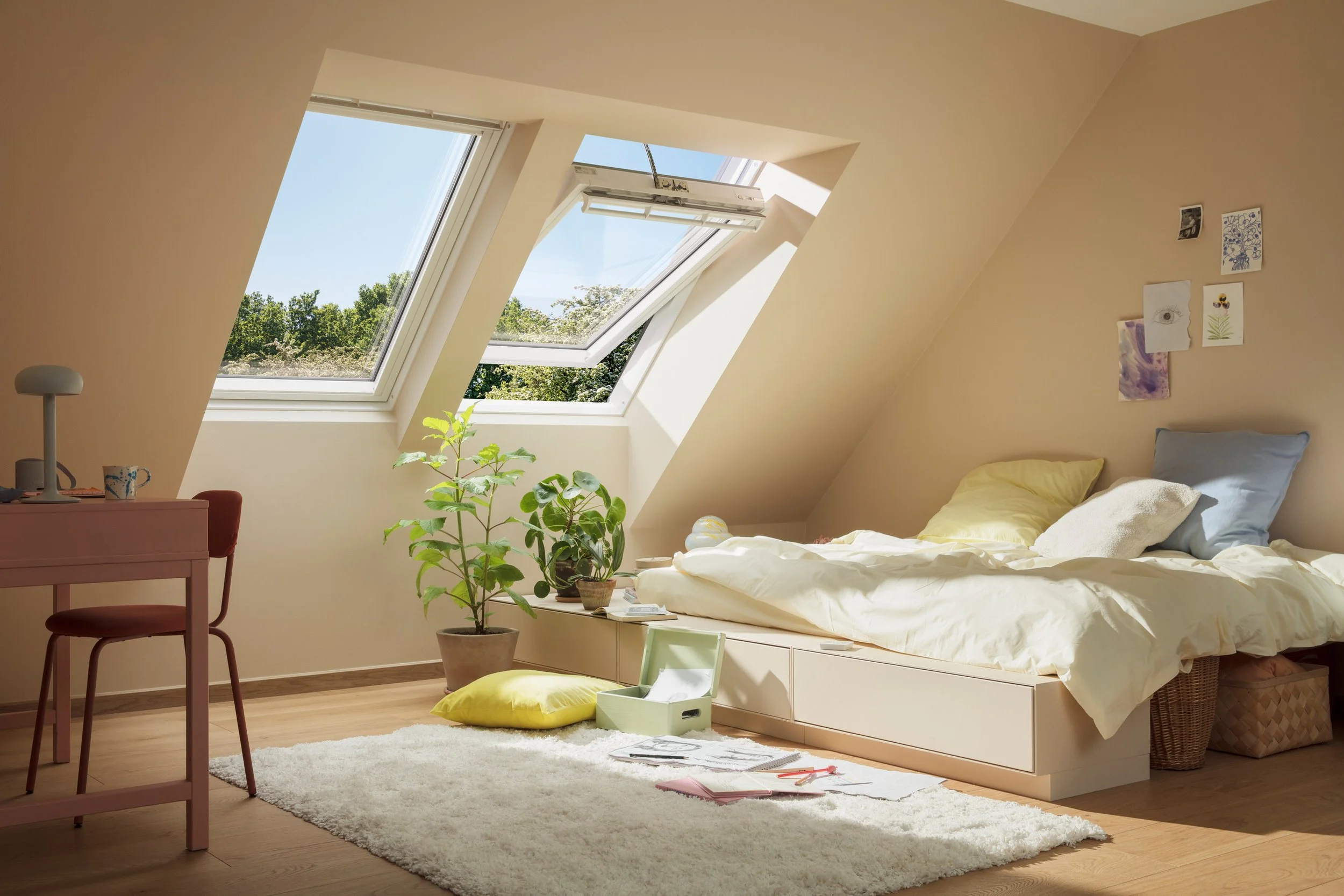 Ein Schlafzimmer mit Dachfenstern, einem ungeordneten Bett mit gelben und blauen Kissen, einem grünen Pflanze, einem kleinen Schreibtisch mit einem weißen Lampen und einer Tasse, und mehreren Bildern an der Wand.