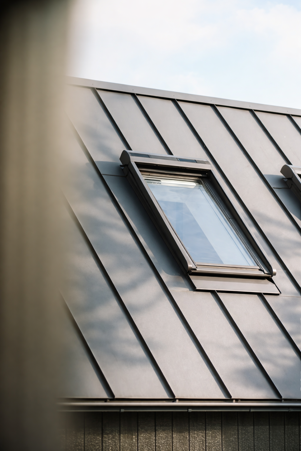 Modernes Hausdach mit neigender Metallverkleidung und einem geöffneten Dachfenster, Blick nach oben auf das Dach und den Himmel.