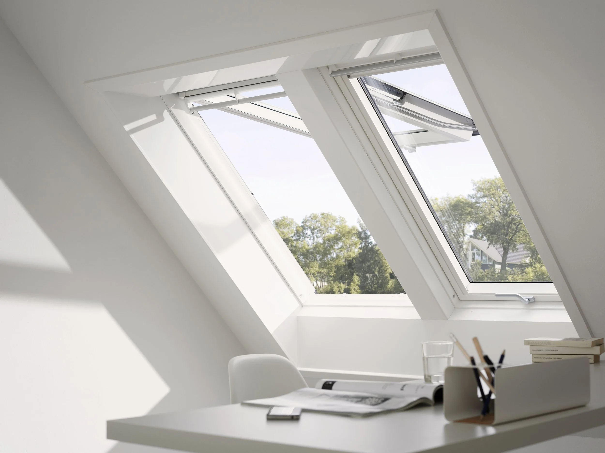 Innenraum mit Dachfenster, hellem weißen Wand und einer weißen Tischseite, auf der Bücher, Schreibutensilien und ein Glas Wasser stehen, bei sonnigem Wetter.