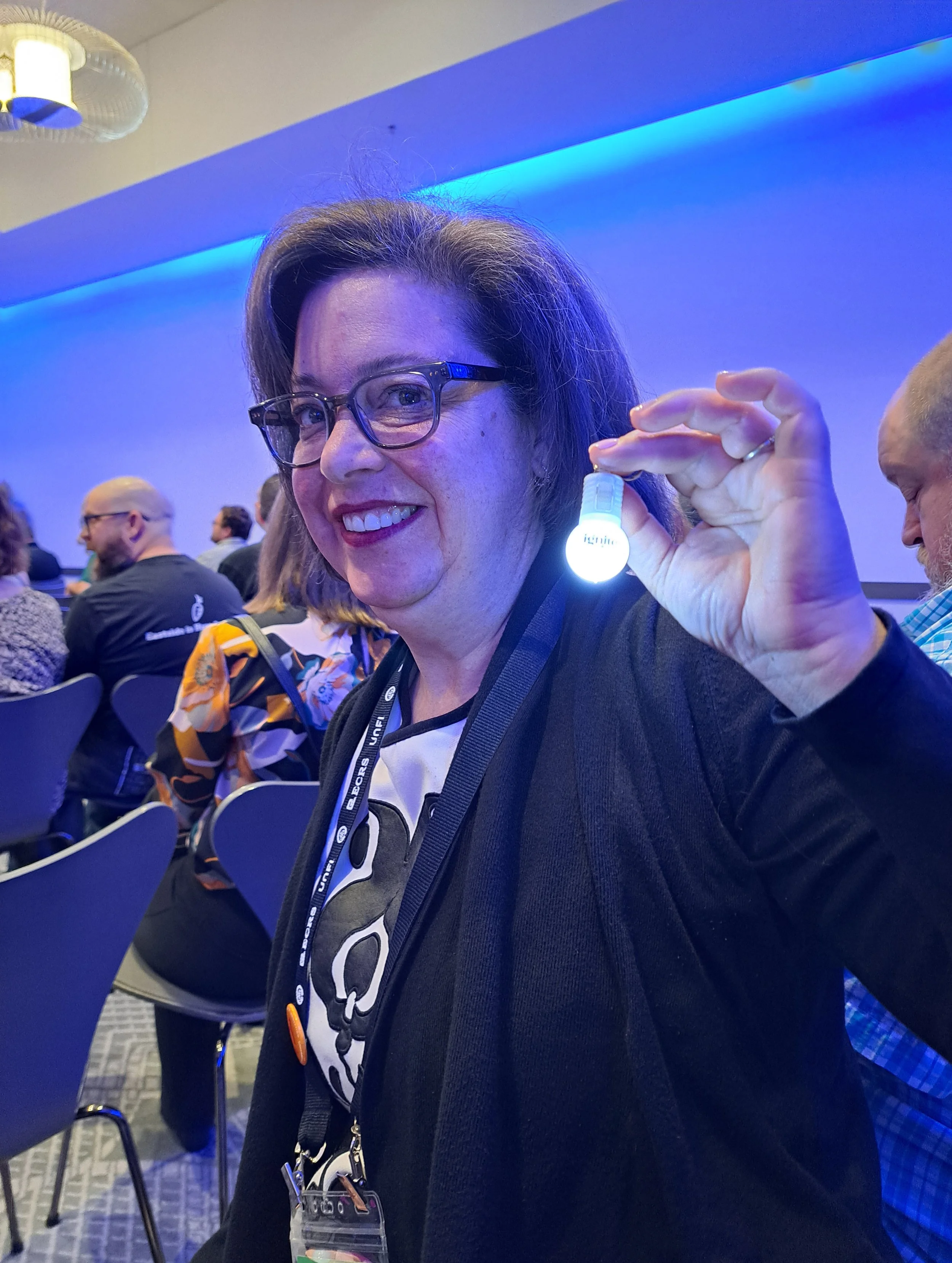 Woman with glasses smiling, holding a small white LED light. Several people are seated in the background in a room with blue lighting.