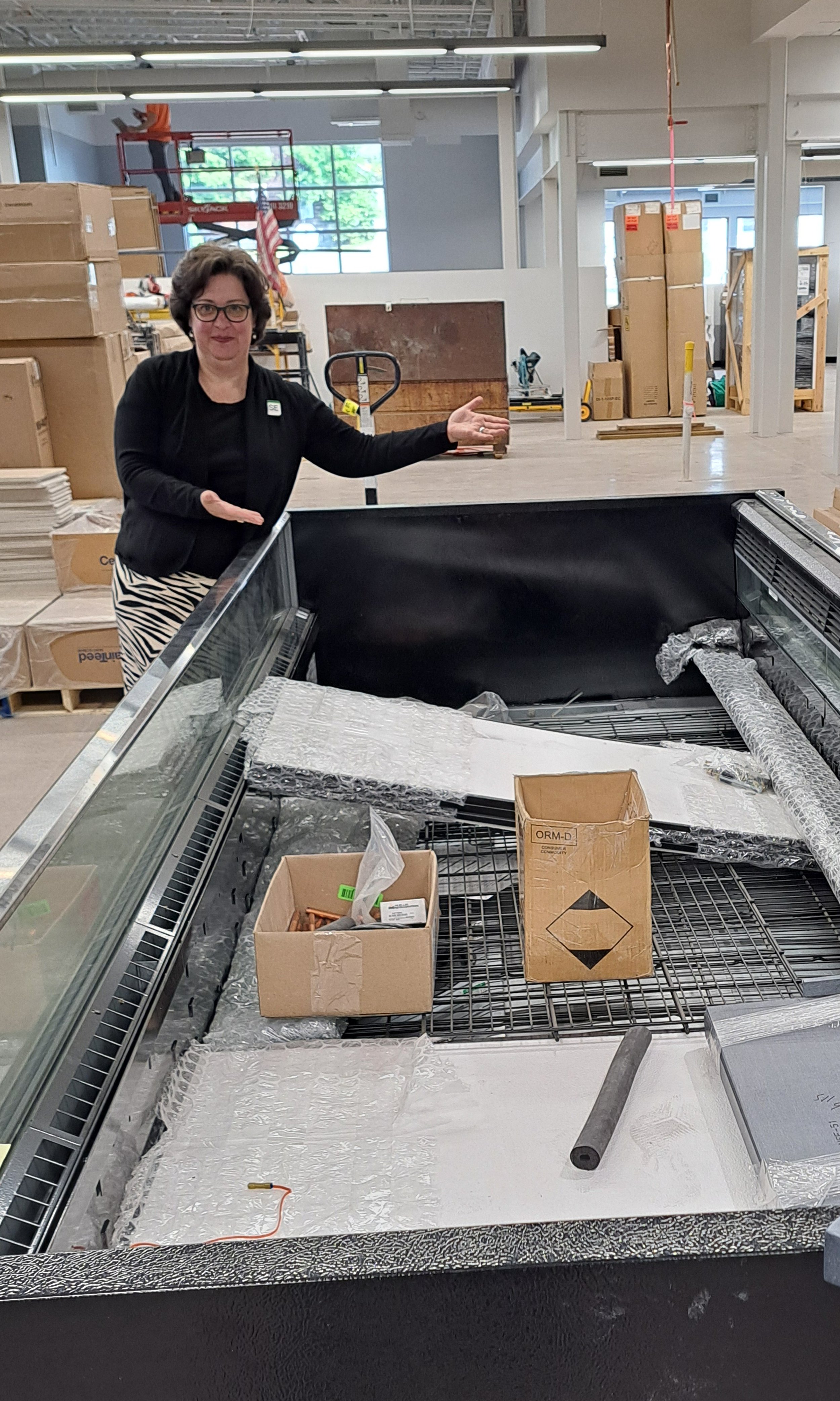 Woman gesturing in an unfinished retail space with empty display cases and boxes, construction materials, and a scissor lift in the background.