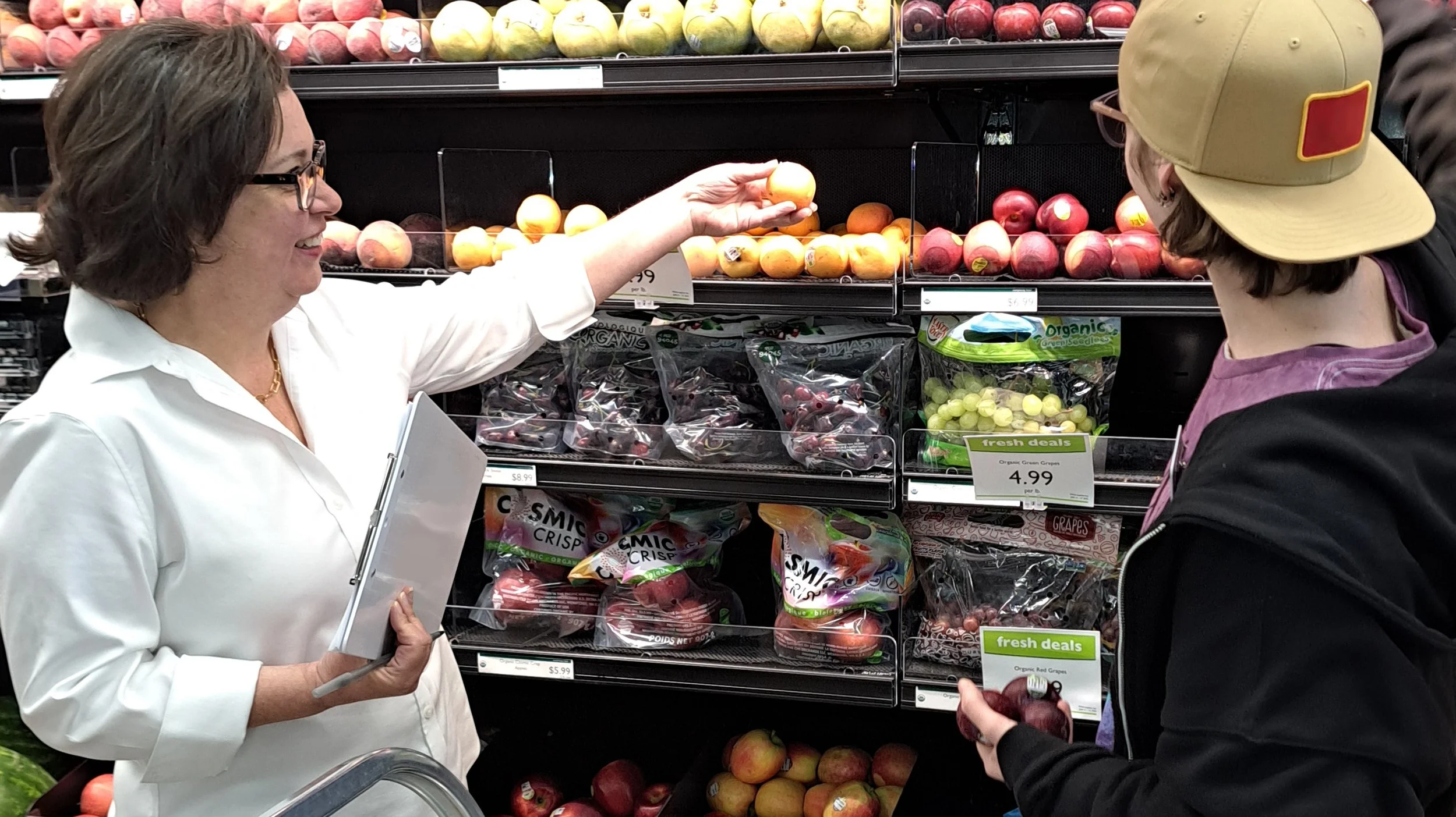 Two women shopping for apples at a grocery store. One woman is handing over a yellow apple to the other. They are standing in front of a produce shelf filled with various apples and grapes.