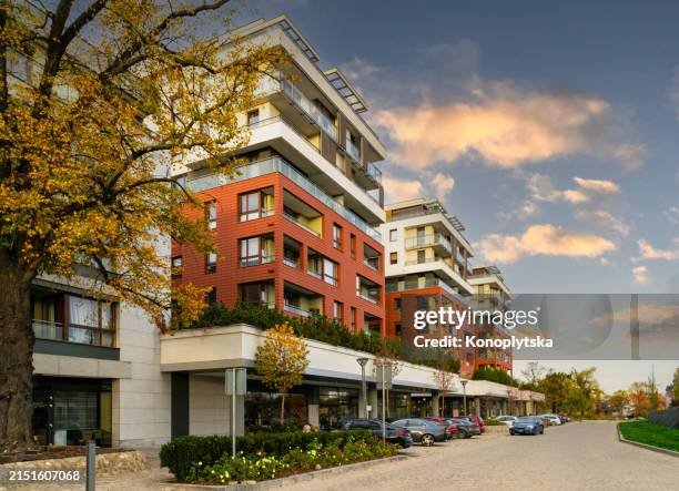 Multi-story modern apartment buildings with red and white facades, parking lot with cars, trees, and a partly cloudy sky.