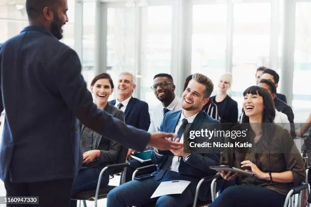 A man stands and hands a business card to a seated woman during a professional presentation or workshop with diverse participants smiling and engaging.