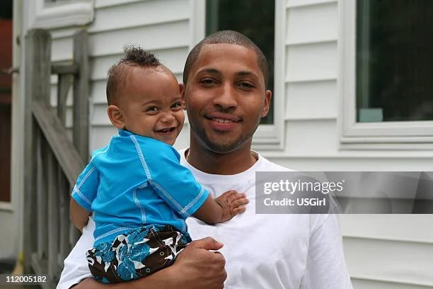 A smiling man holding a young boy outside in front of a white house.