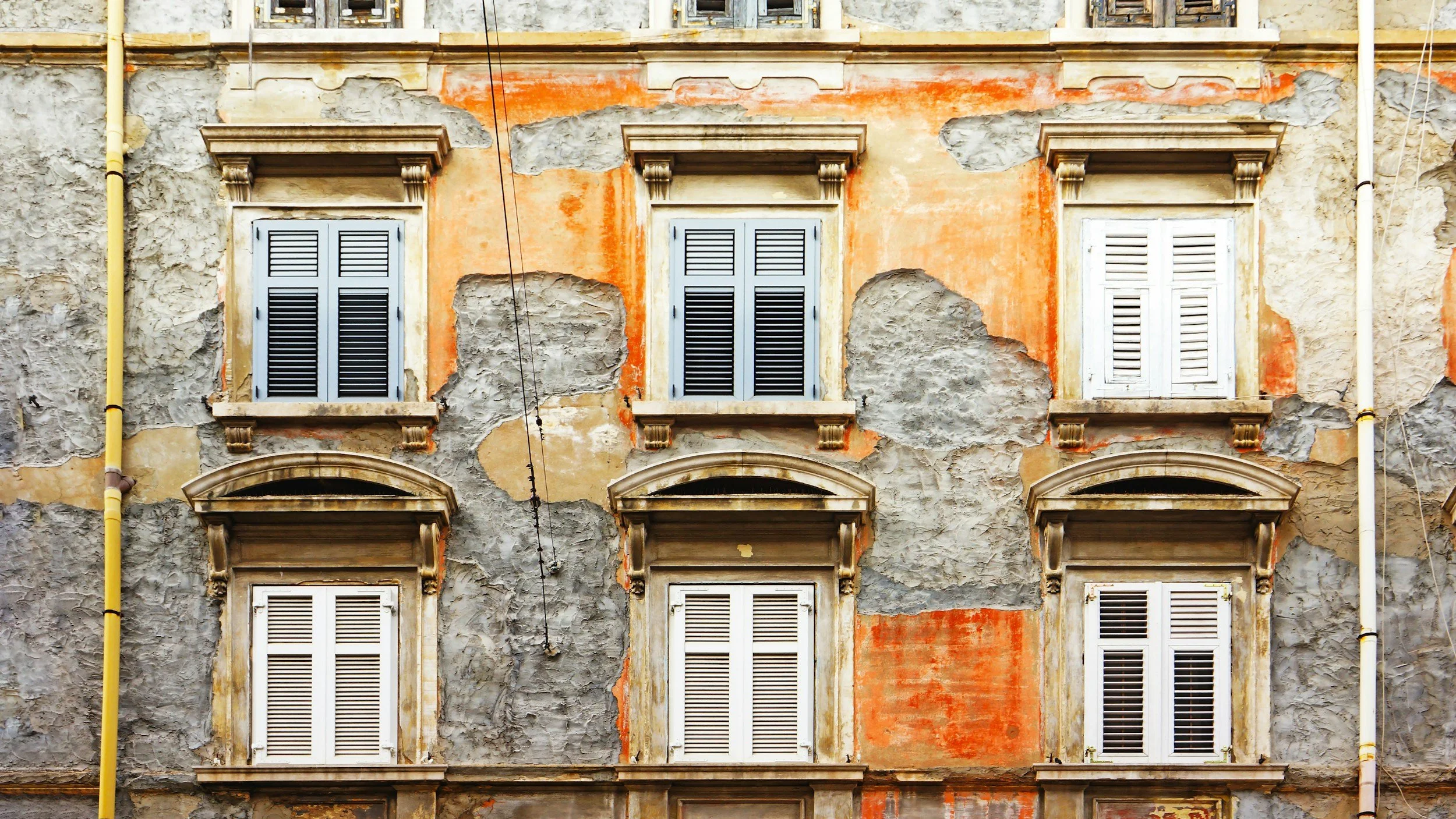 Old building facade with six windows with shutters, some shutters closed, and weathered, peeling walls with patches of orange and gray paint.