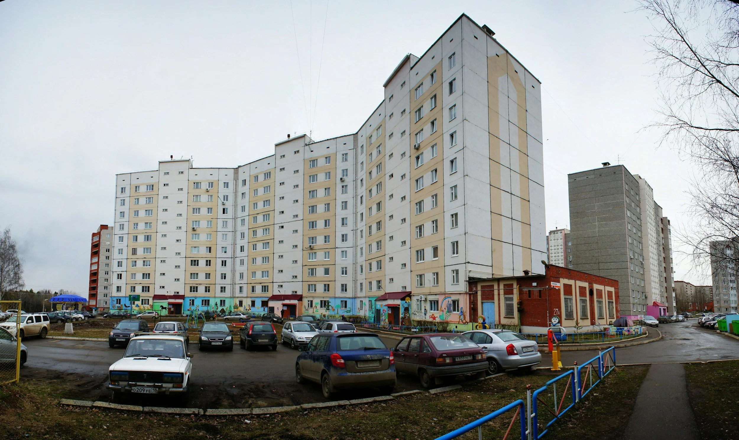 A residential area with multiple high-rise apartment buildings, a parking lot with cars, and a small playground with colorful equipment, under an overcast sky.
