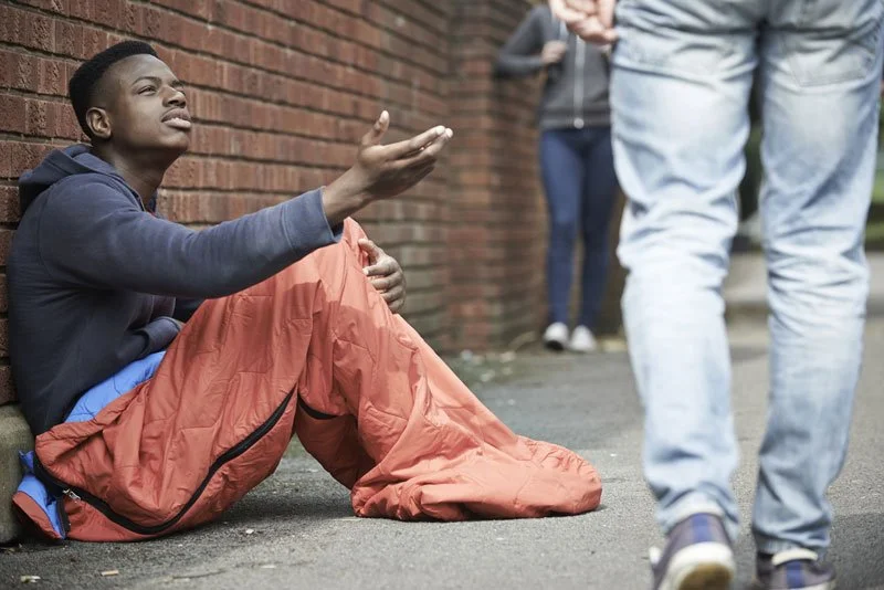 A young man sitting on the sidewalk, reaching out with one hand, talking to a person standing nearby, with others in the background near a brick wall.