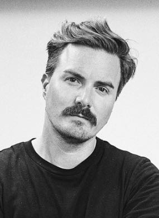 Black and white portrait of a man with styled hair and a mustache, wearing a dark shirt.