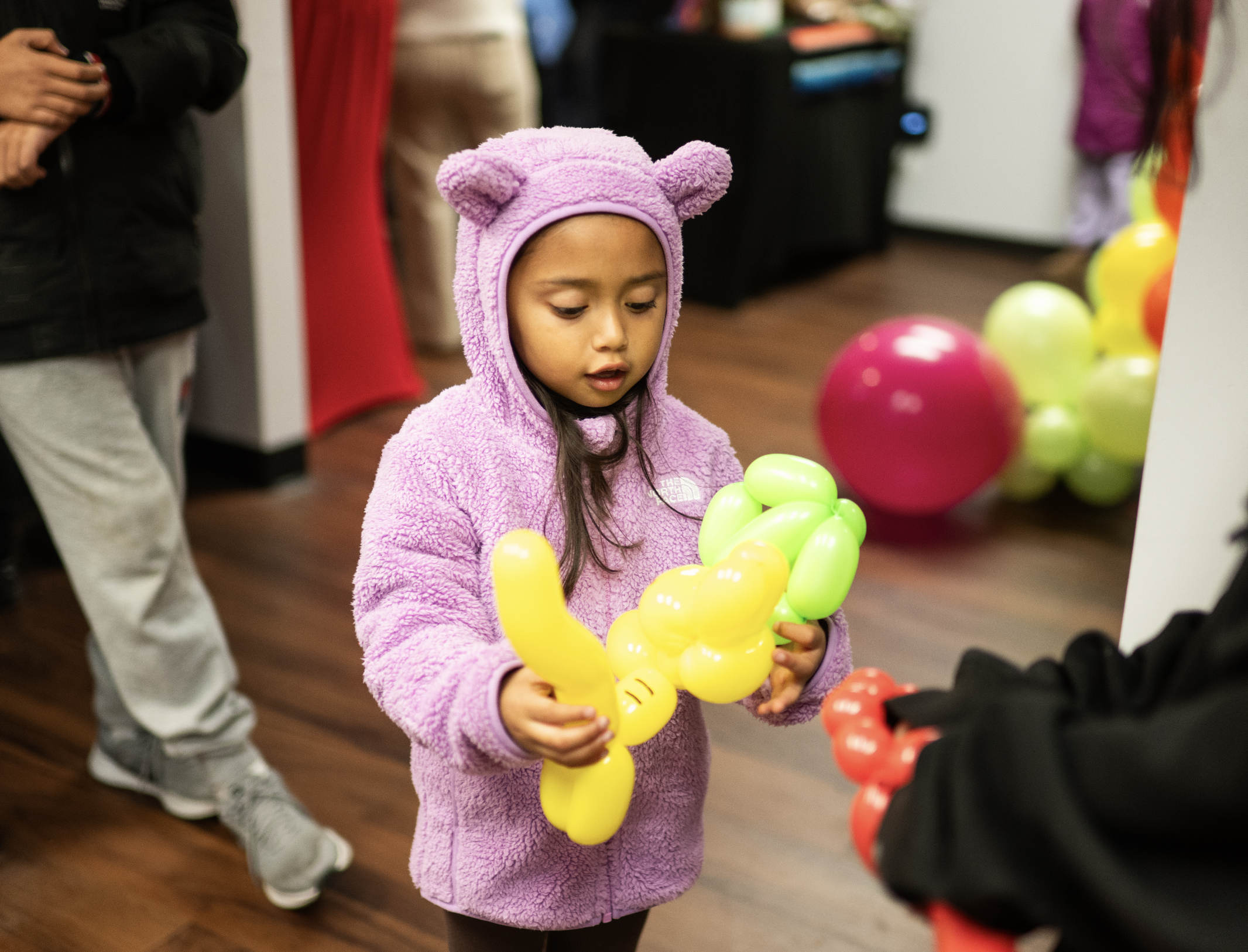 A young girl wearing a pink bear hoodie is holding a yellow balloon animal at an indoor event. Others are nearby, and colorful balloons are visible in the background.