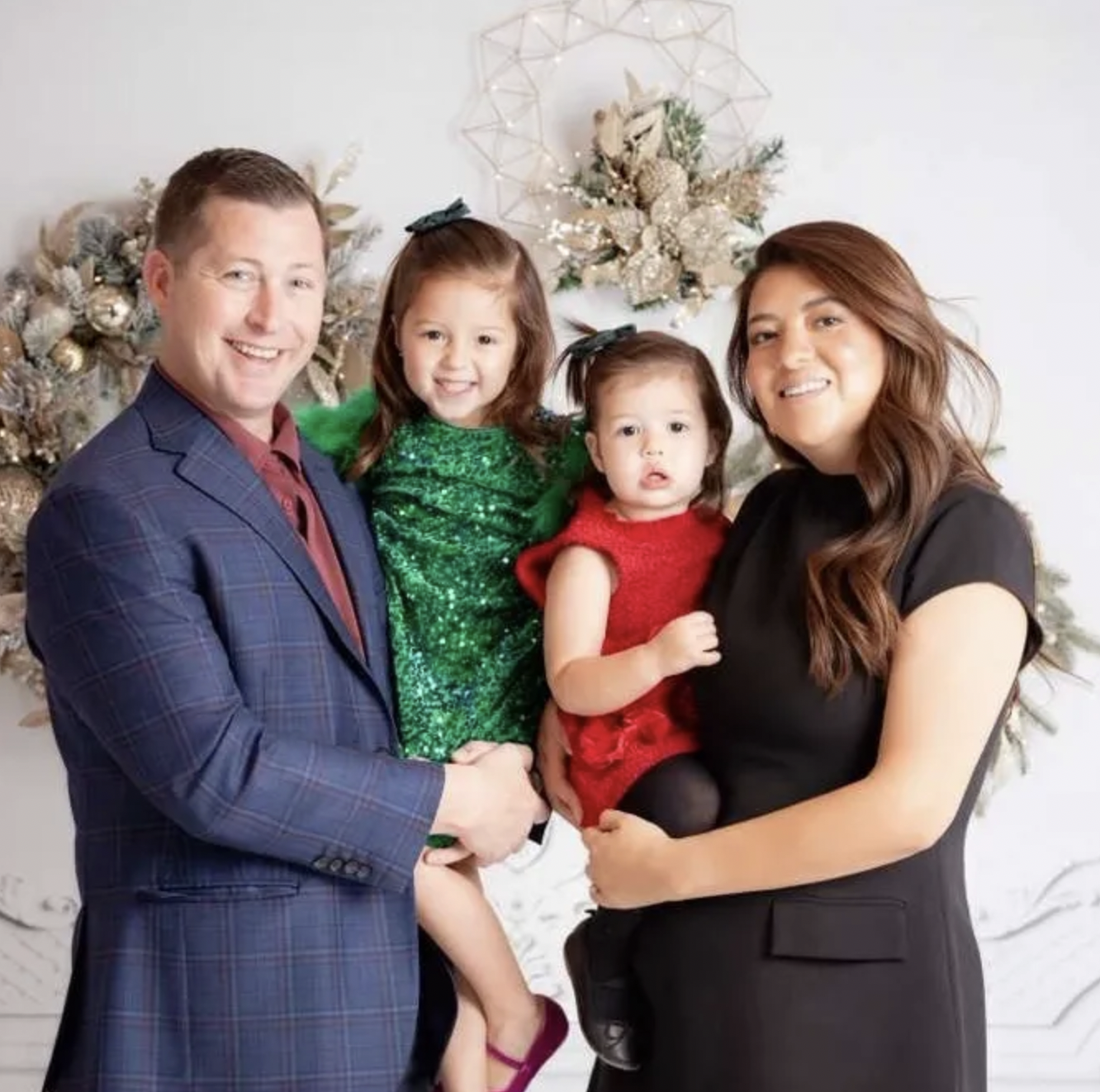 A smiling family of four posing in front of Christmas decorations, including a Christmas tree with ornaments and a decorative wreath or star on the wall behind them.
