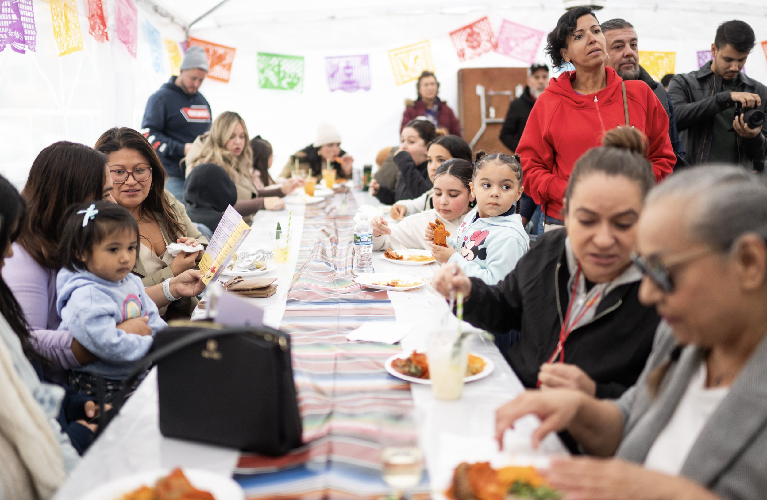 People seated at a long table with plates of food and drinks at a festive gathering under colorful papel picado banners.