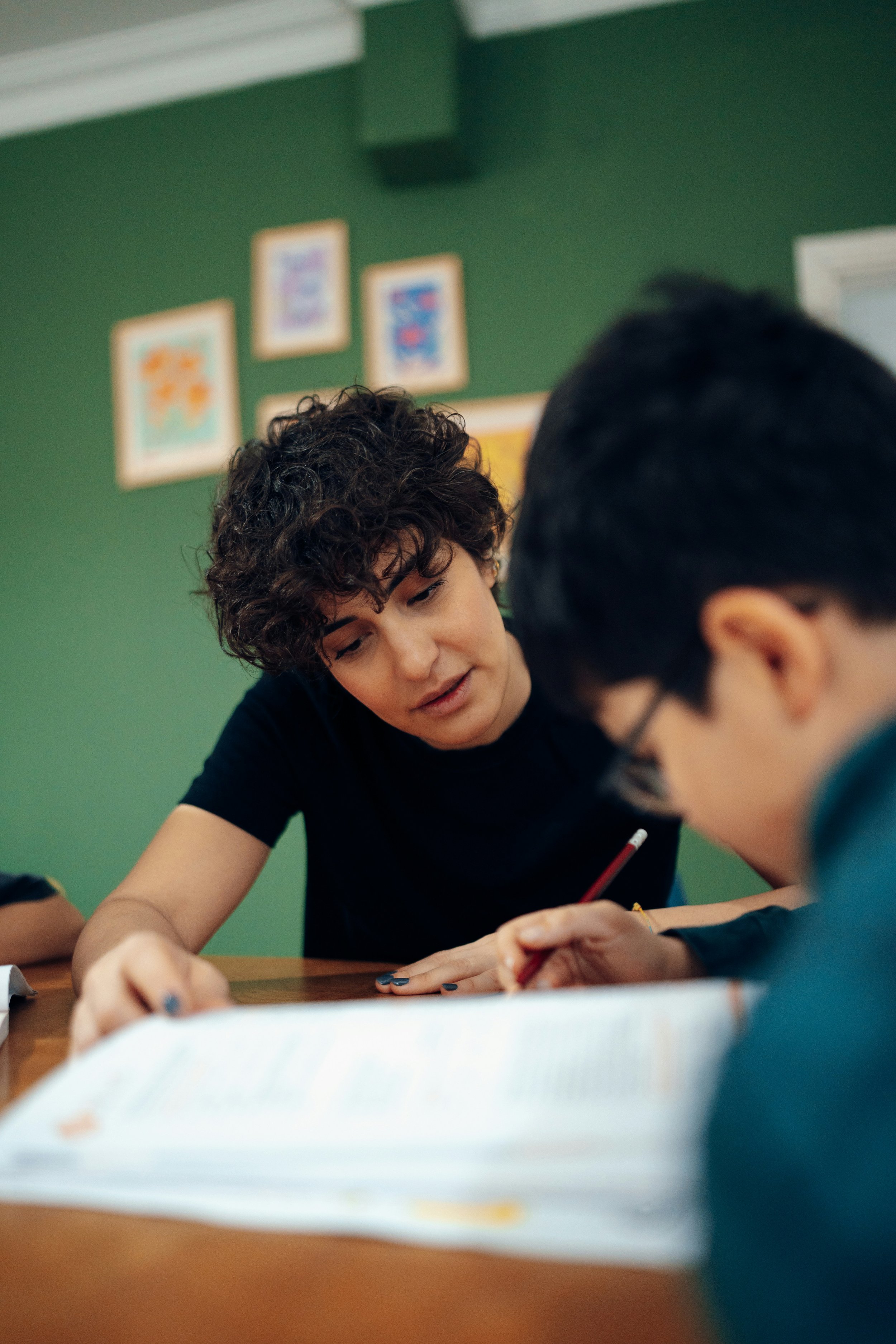A woman with curly dark hair leaning over a table, assisting a young man with glasses and dark hair who is writing with a red pencil, in a room with green walls and framed artwork.