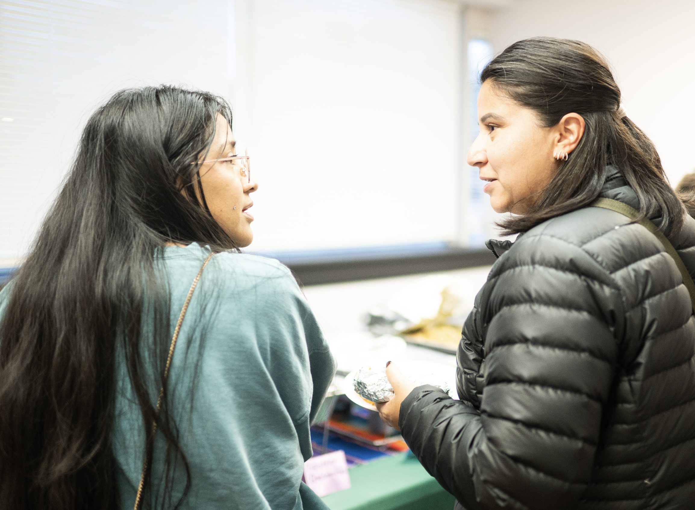 Two women are facing each other closely indoors. One woman with long dark hair and glasses wears a teal jacket, and the other woman with dark hair in a ponytail and earrings wears a black puffer jacket. They appear to be having a conversation or sharing a moment.