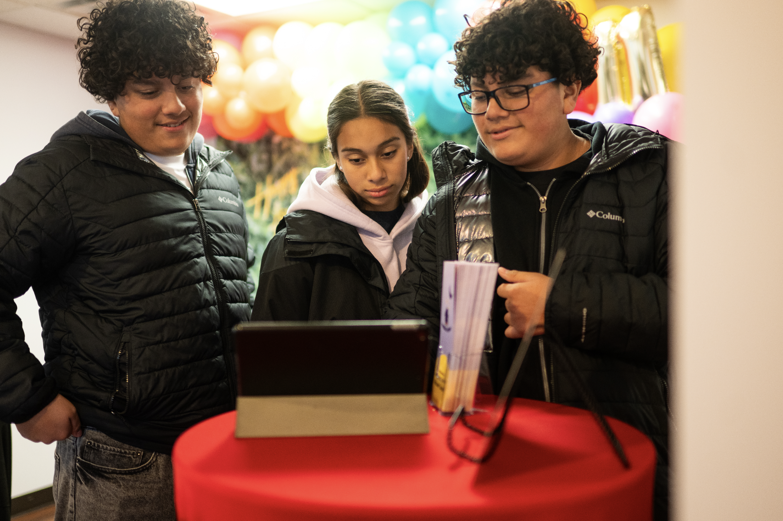 Three children, two boys and a girl, are gathered around a table at an indoor event with colorful balloons in the background. They are looking at something on a device or paper, with a red table in front of them.