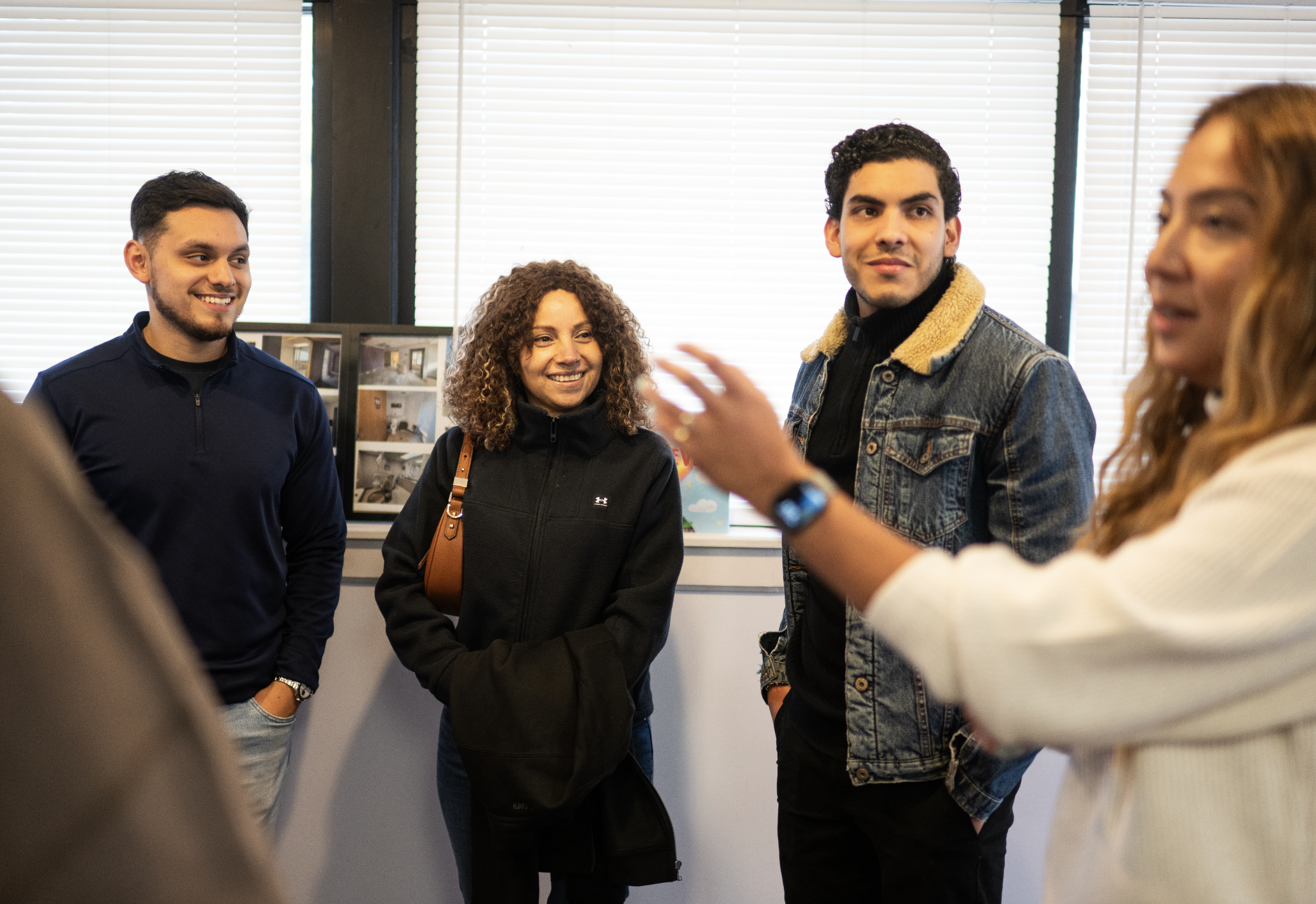 Four people engaged in a conversation in an office setting, with blinds covering the windows in the background.