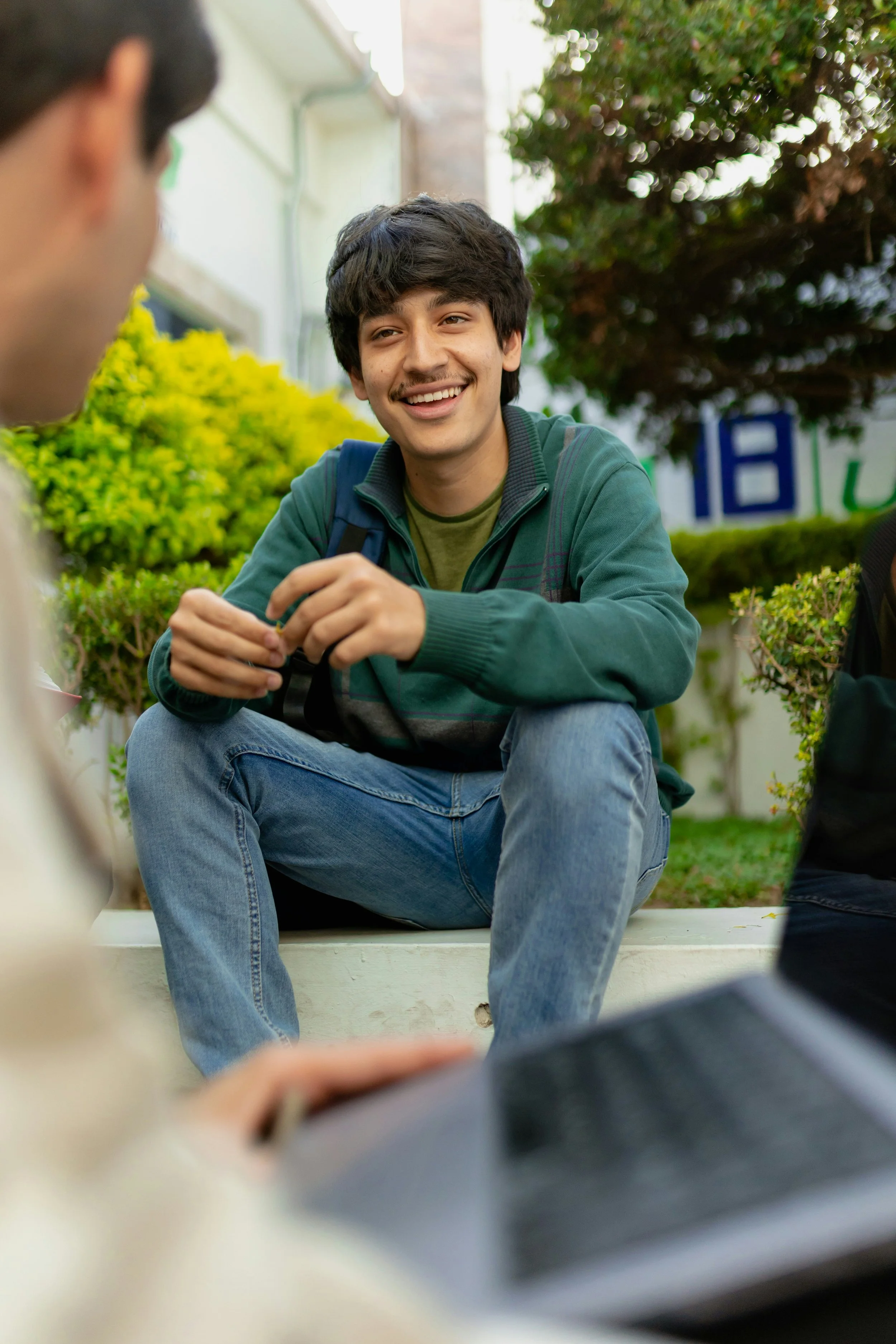 A young man with dark hair and a mustache sitting outdoors on a bench, smiling and engaging in conversation with another person
