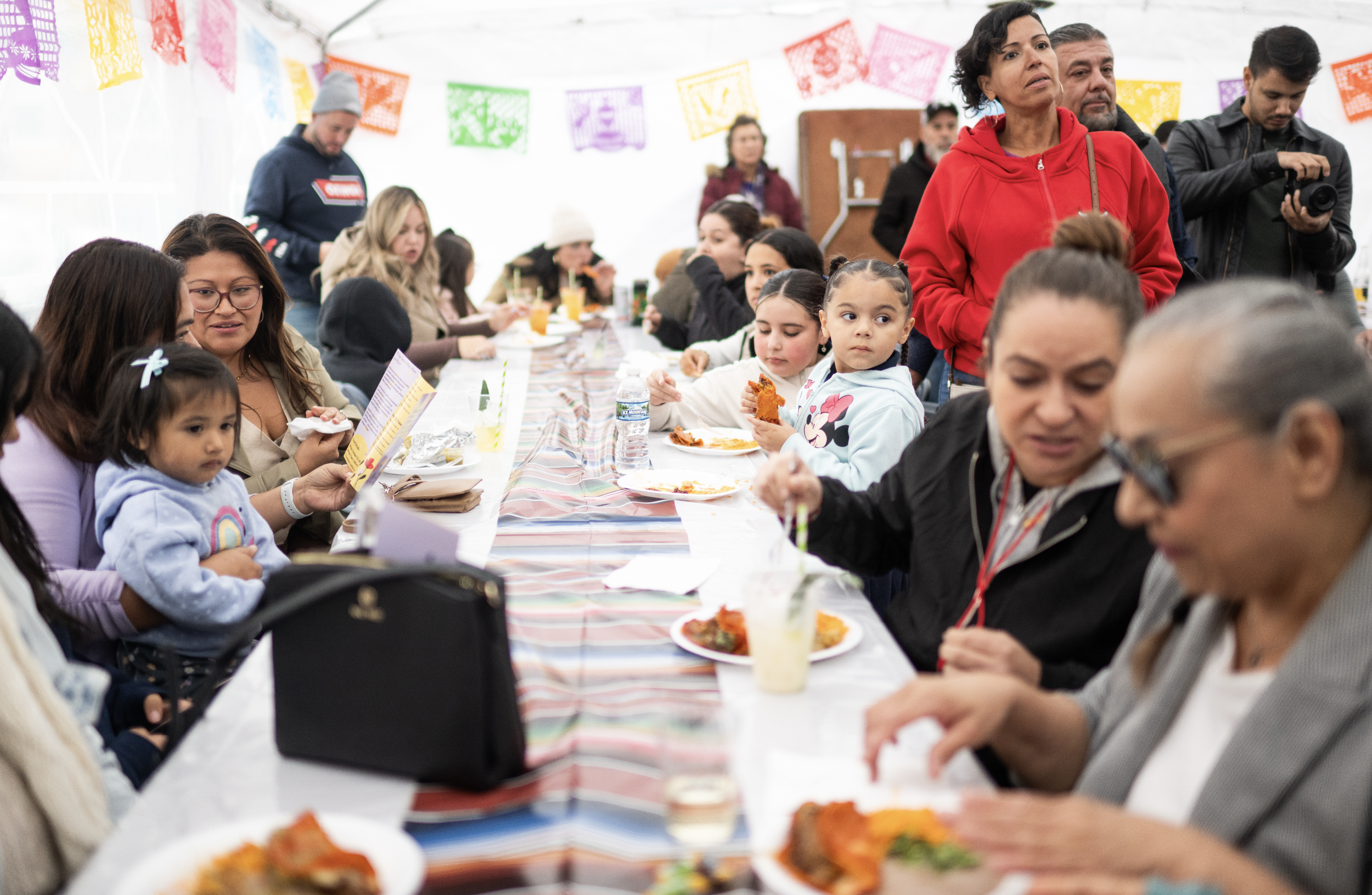 A large group of people, including children and adults, sitting at a long table under a tent decorated with colorful papel picado banners, enjoying a meal at a festive gathering.