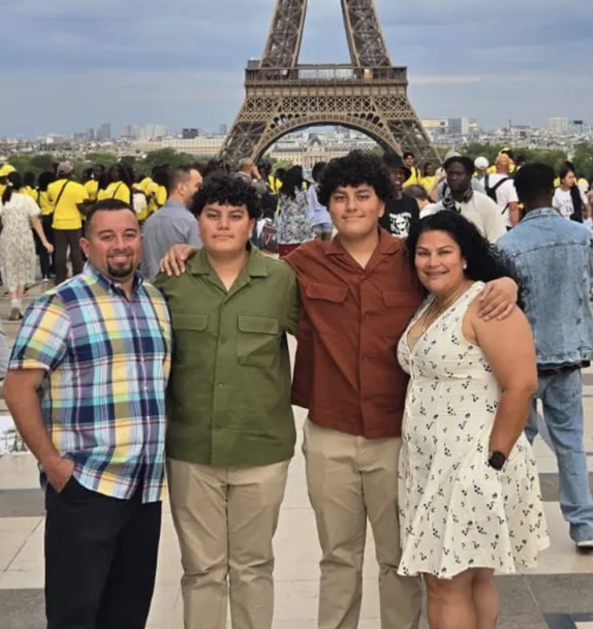 Family of four posing for a photo in front of the Eiffel Tower in Paris, France, with a crowd of tourists in the background.