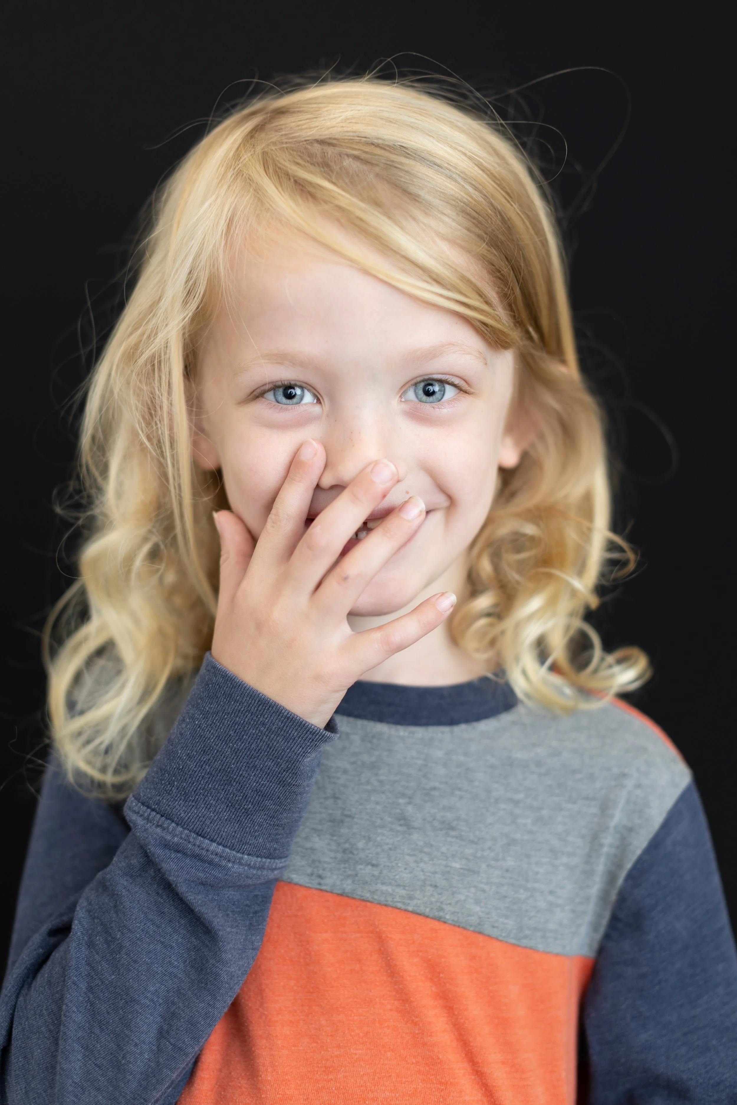 Young girl with long blonde hair and blue eyes, smiling and covering her mouth with her hand, wearing a gray and orange shirt, against a black background.