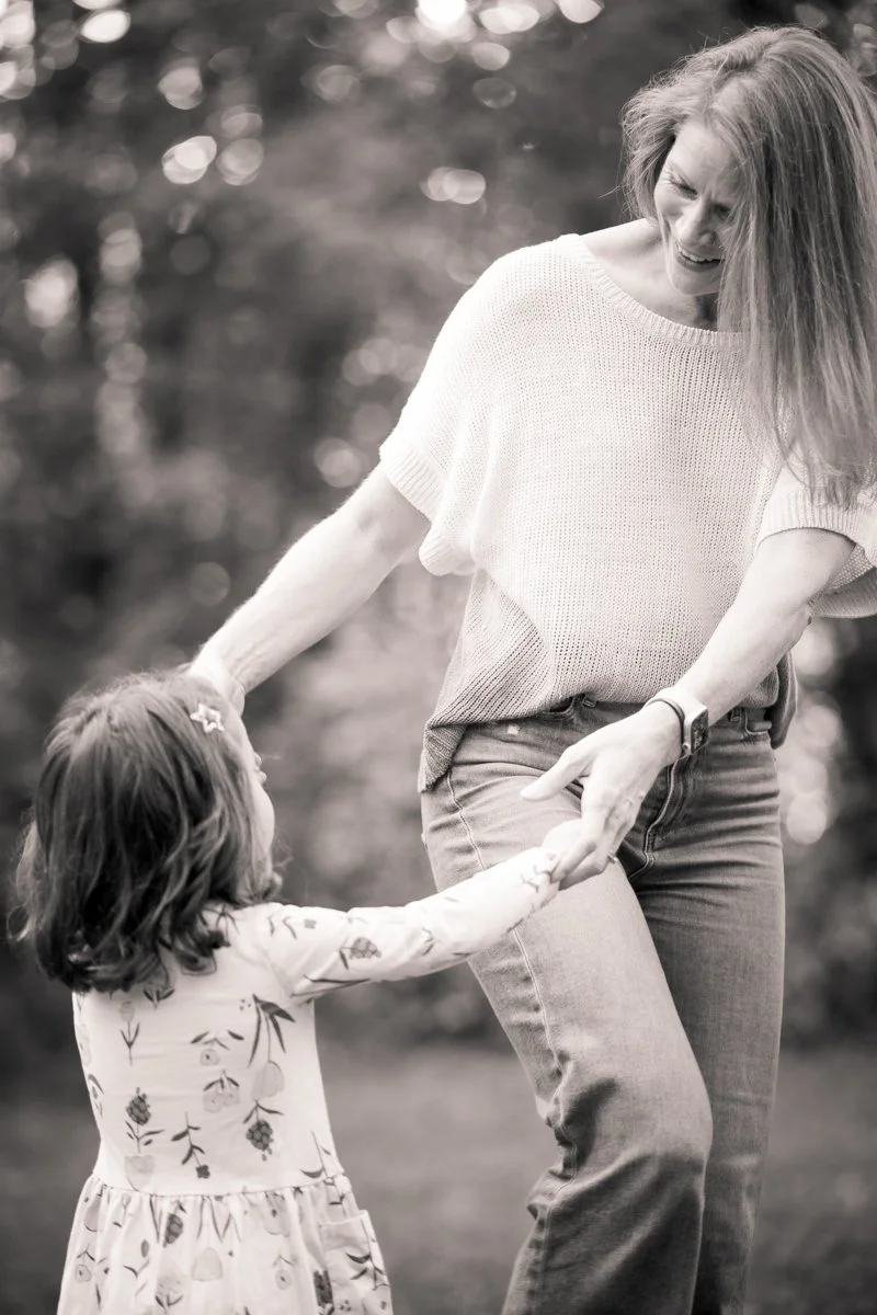 A woman and a young girl holding hands outdoors. The woman is smiling and looking down at the girl, who is looking up at her. The woman has long hair and is wearing a light-colored sweater and jeans, while the girl has shoulder-length hair and is wearing a floral dress.