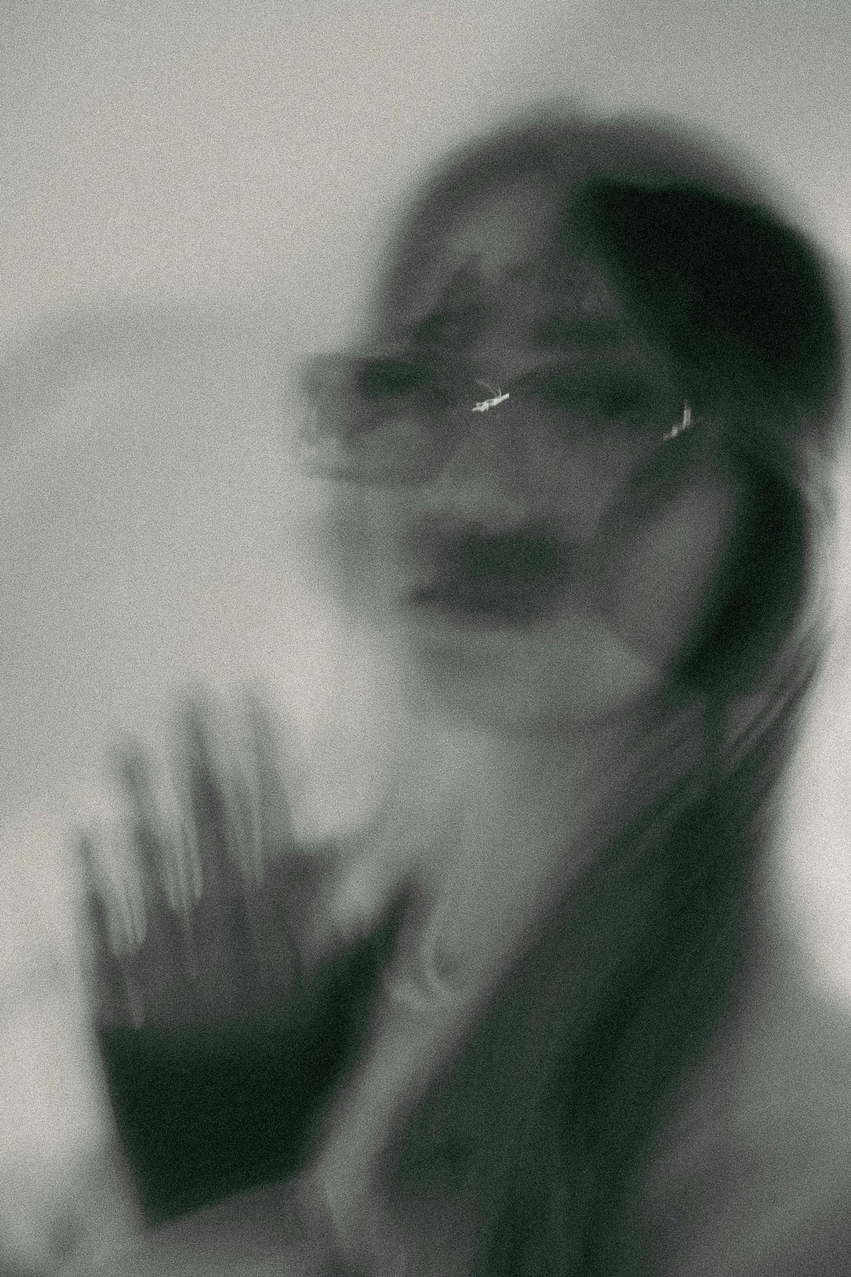 A blurry black-and-white photo of a woman with sunglasses, dark lipstick, and long hair, holding her hand up near her face.