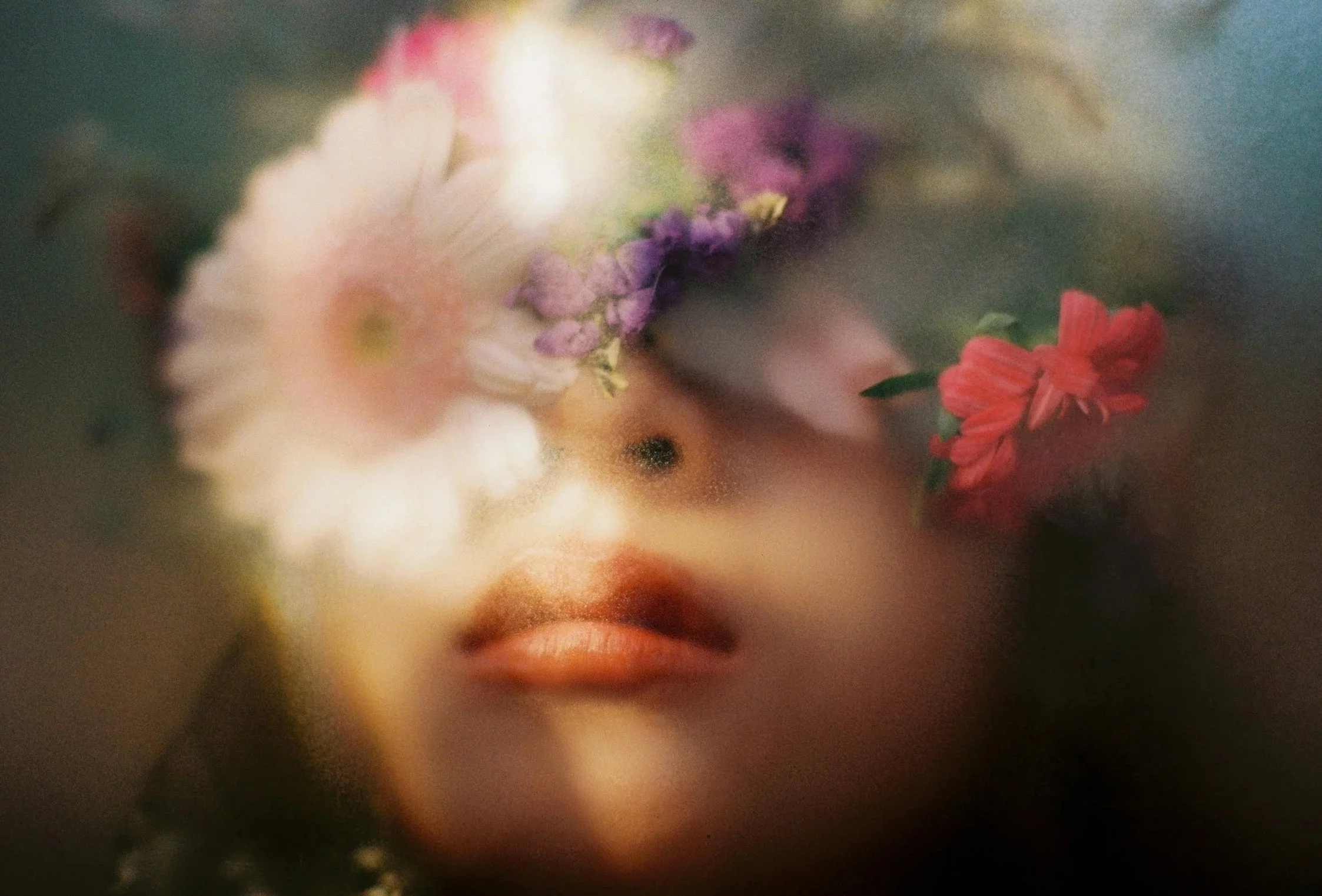 A blurred, artistic close-up of a woman's face with closed lips, surrounded by pastel-colored flowers.