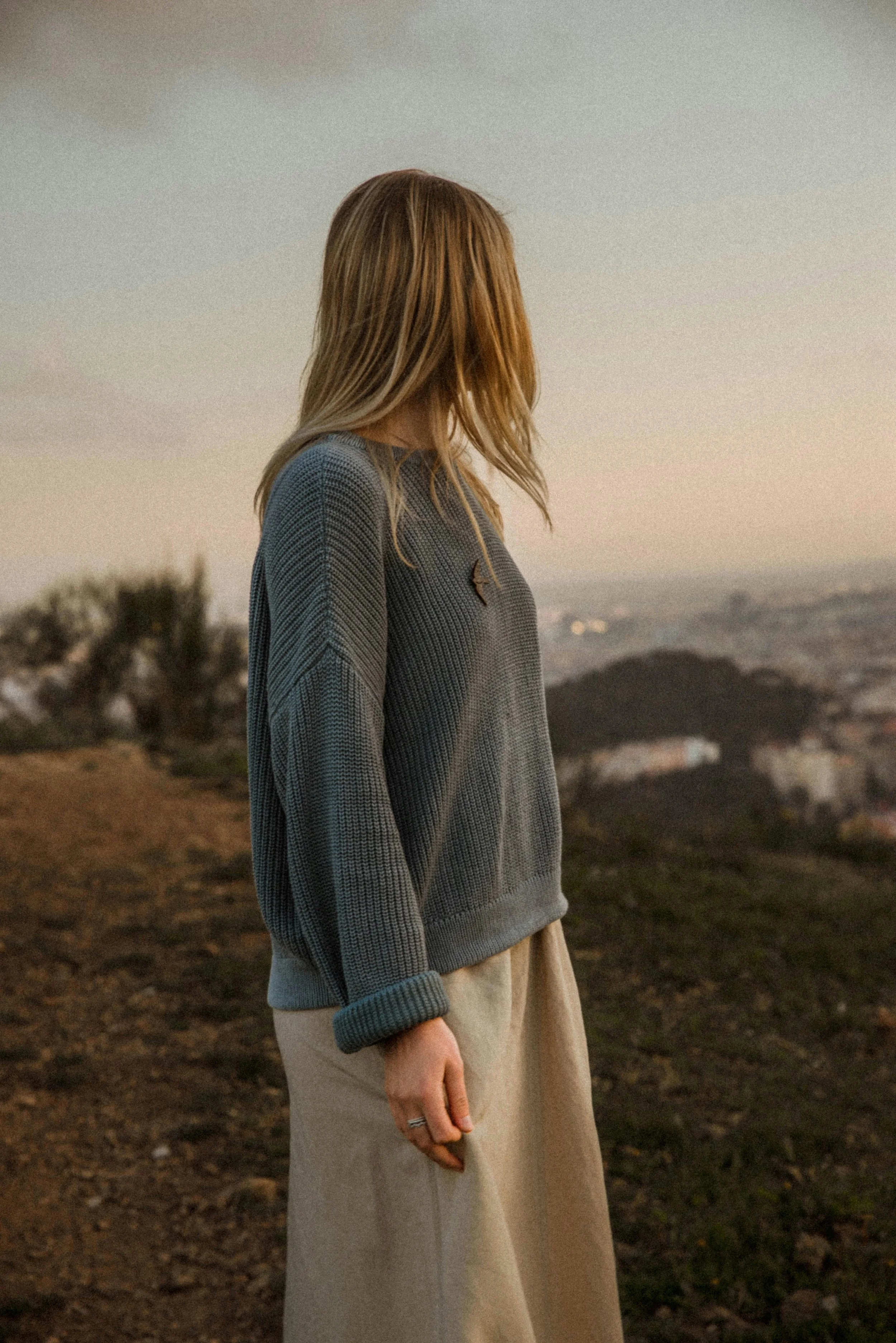 A woman standing outdoors on a hill during sunset, with her side to the camera, wearing a gray sweater and beige skirt, looking towards the horizon.